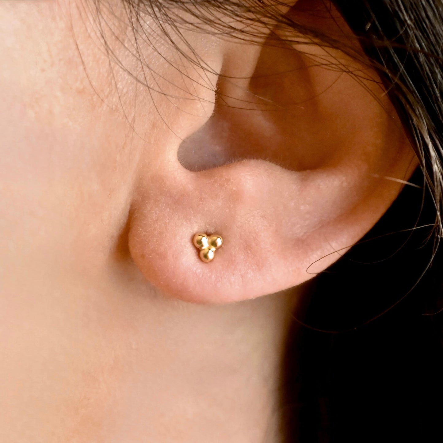 A close-up of a person's ear wearing a small gold stud earring with three clustered balls.