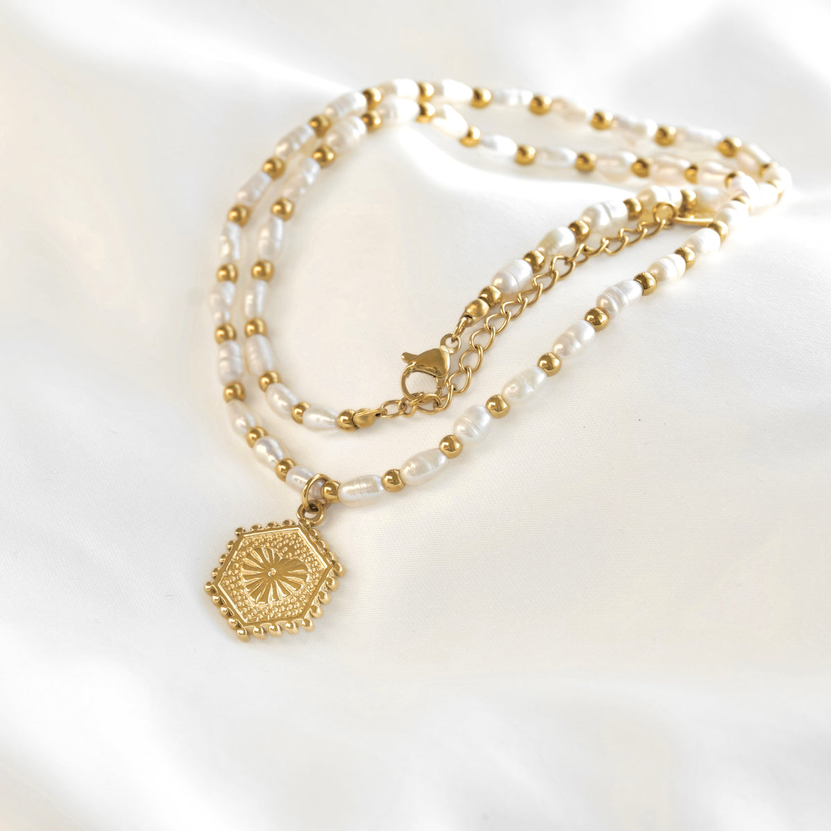 A delicate double-strand necklace featuring white freshwater pearls and small gold beads, with a gold chain and a hexagonal gold pendant, laid out on a white satin background.