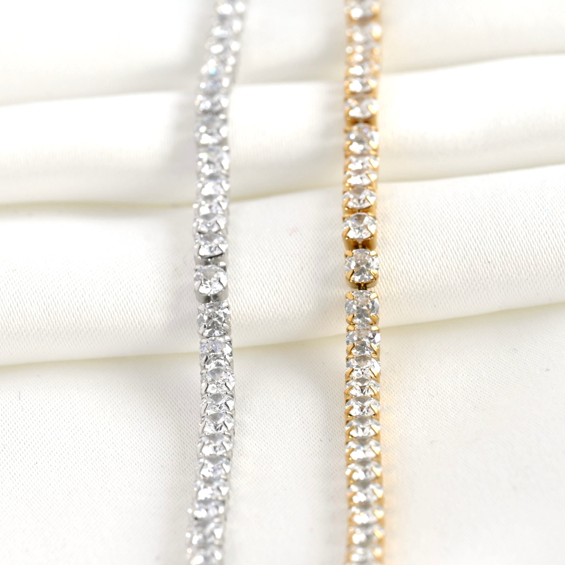 Front vertical view of two diamond tennis chains, one silver and one gold, on a white fabric background.