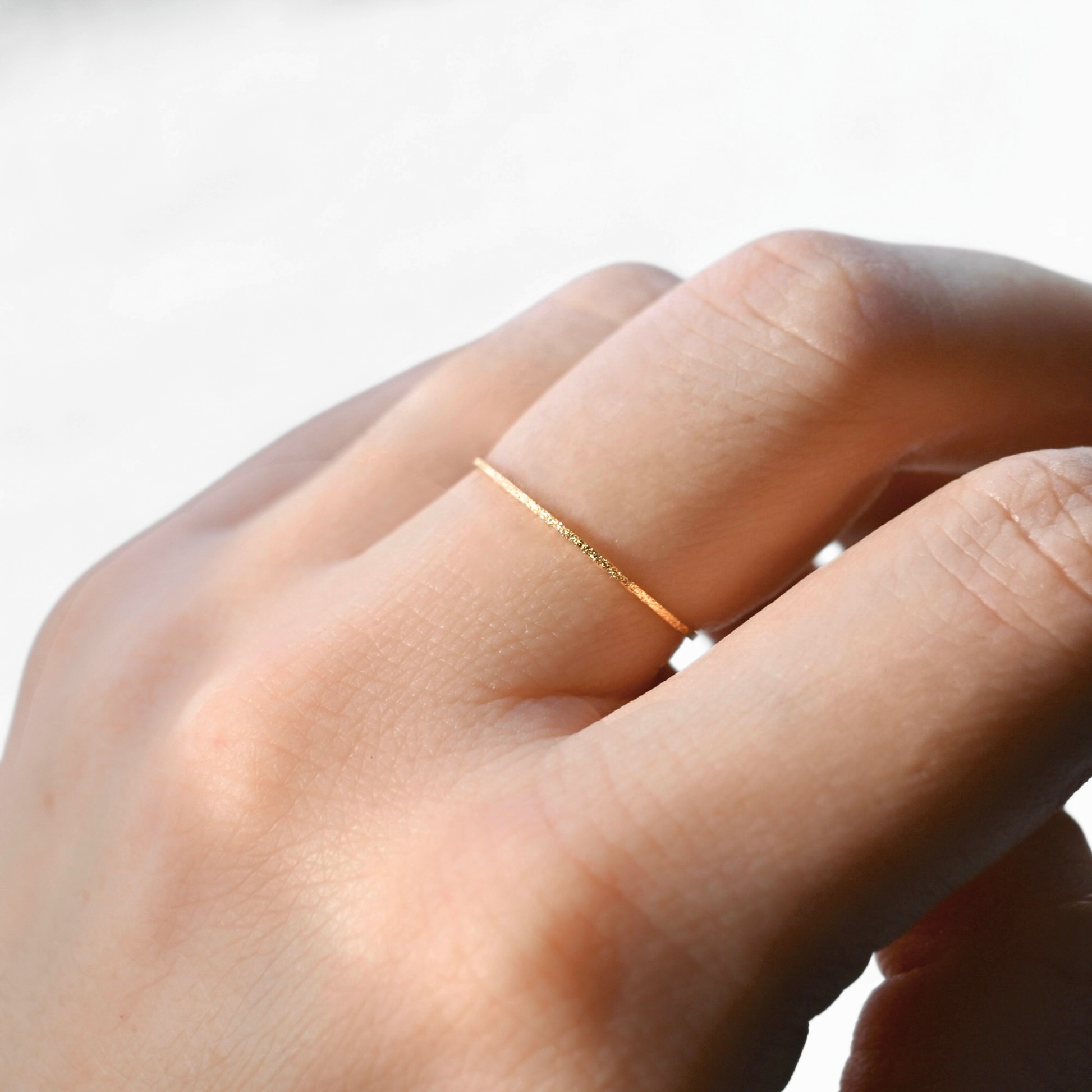 A close-up of a hand wearing a very thin, delicate gold ring on one of the fingers against a bright, light background.