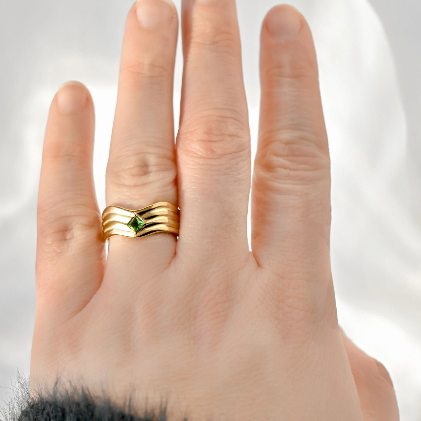 A close-up of a hand wearing a gold ring with three wavy bands and a small green diamond-shaped gemstone in the center, set against a soft white background.