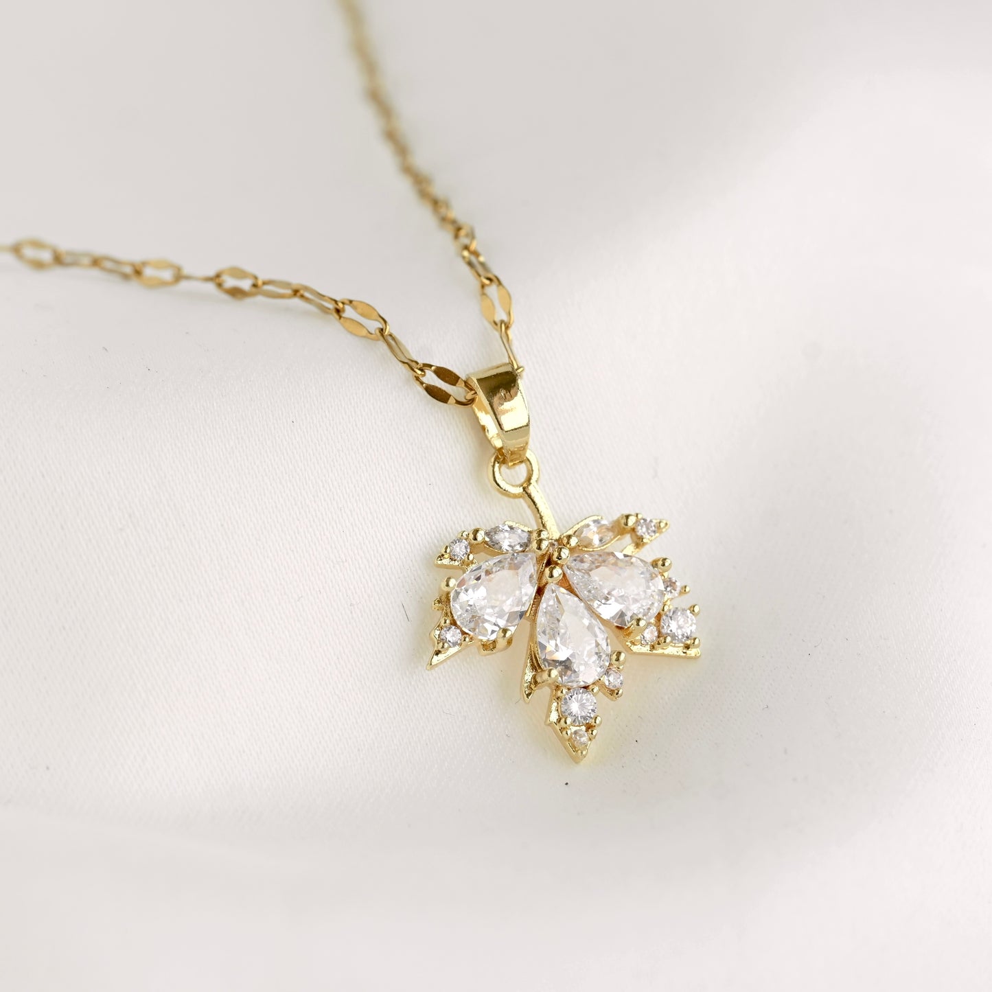 A gold pendant necklace featuring a maple leaf set with clear teardrop and round gemstones, displayed on a soft white fabric background.