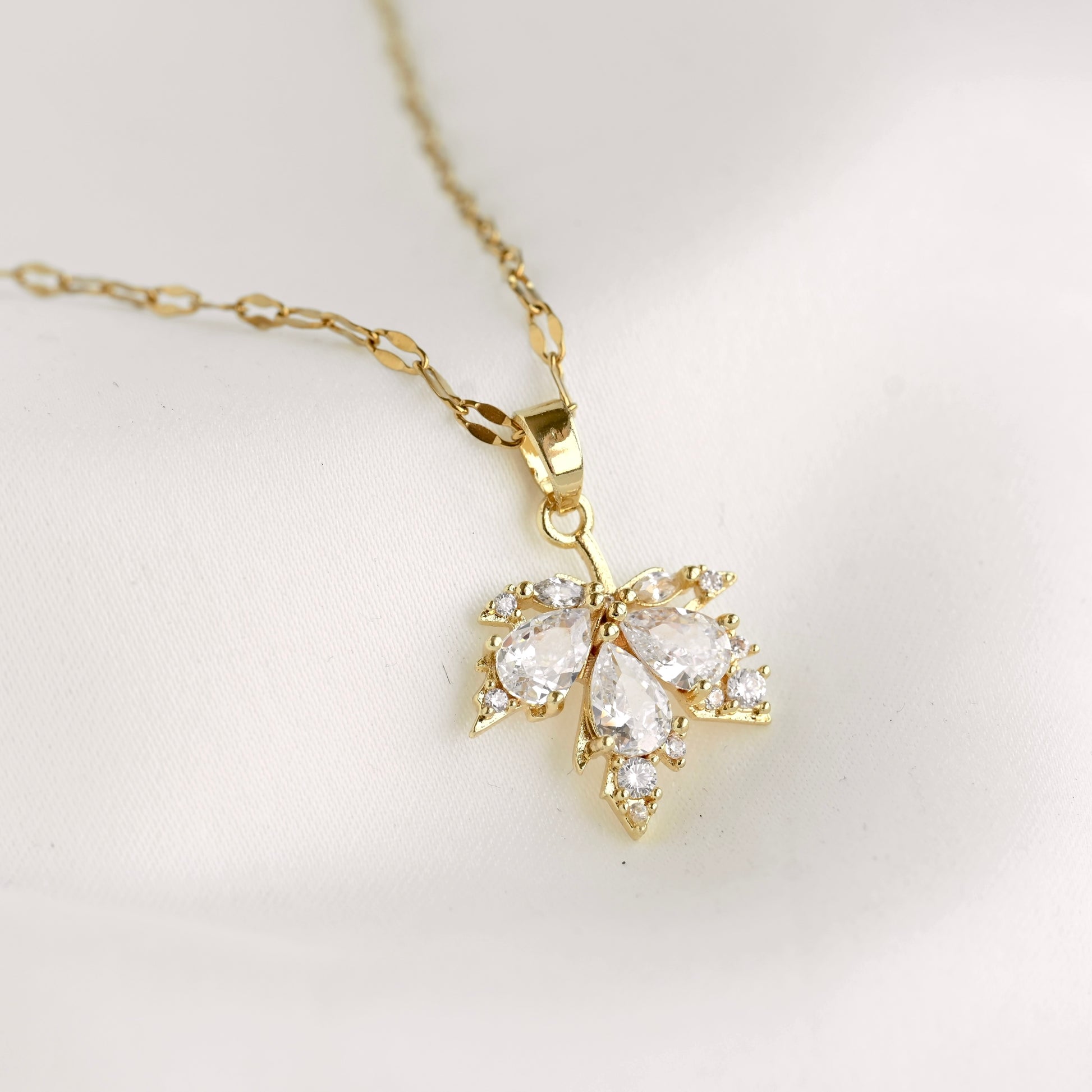 A gold pendant necklace featuring a maple leaf set with clear teardrop and round gemstones, displayed on a soft white fabric background.