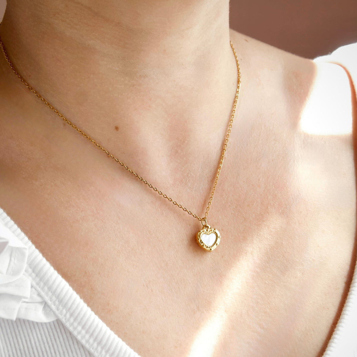 A delicate gold chain necklace with a small heart-shaped pendant featuring a white inlay, worn on a model's neck over a white top.