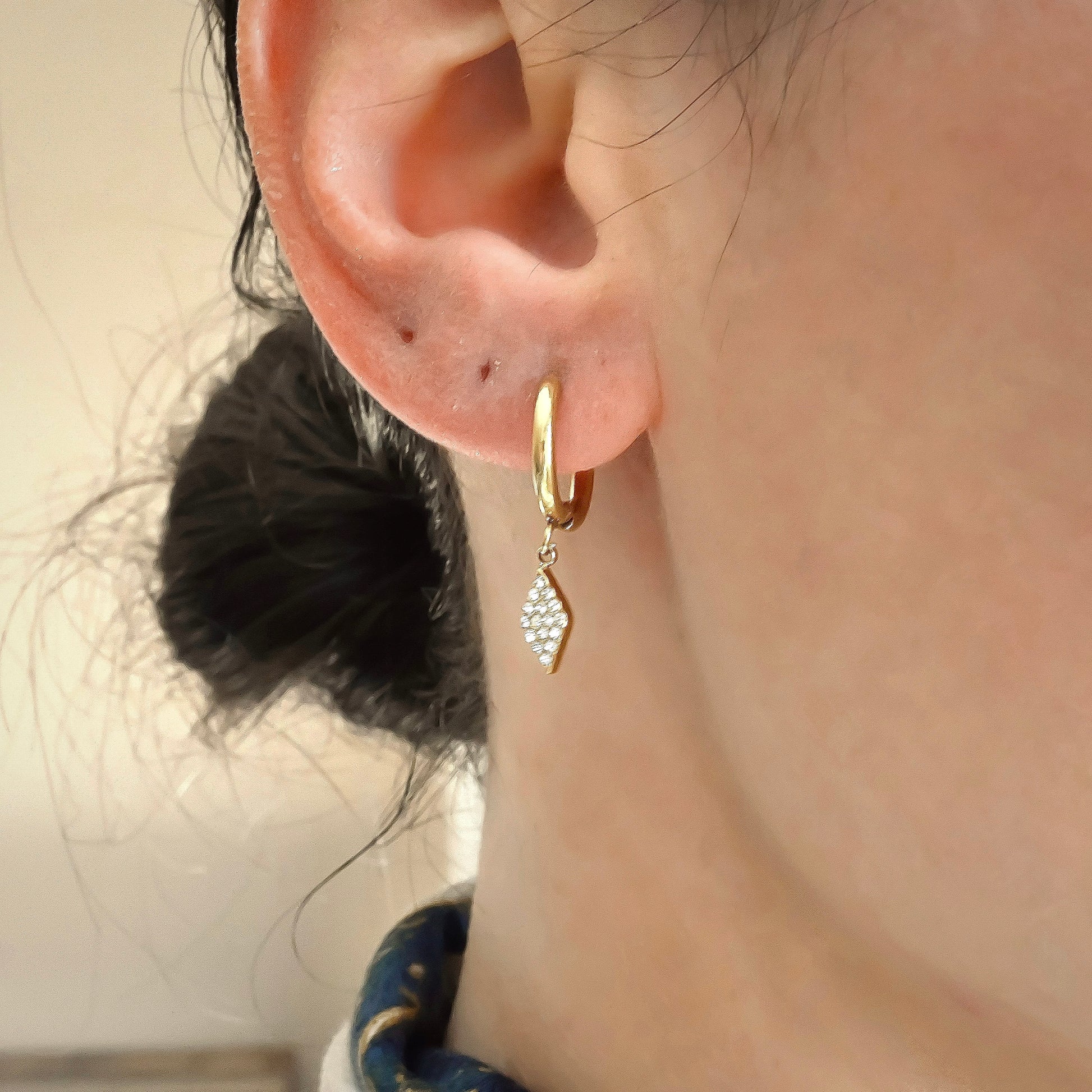 Gold huggie hoops with rhombus dangles pave set with clear stones on a person's ear