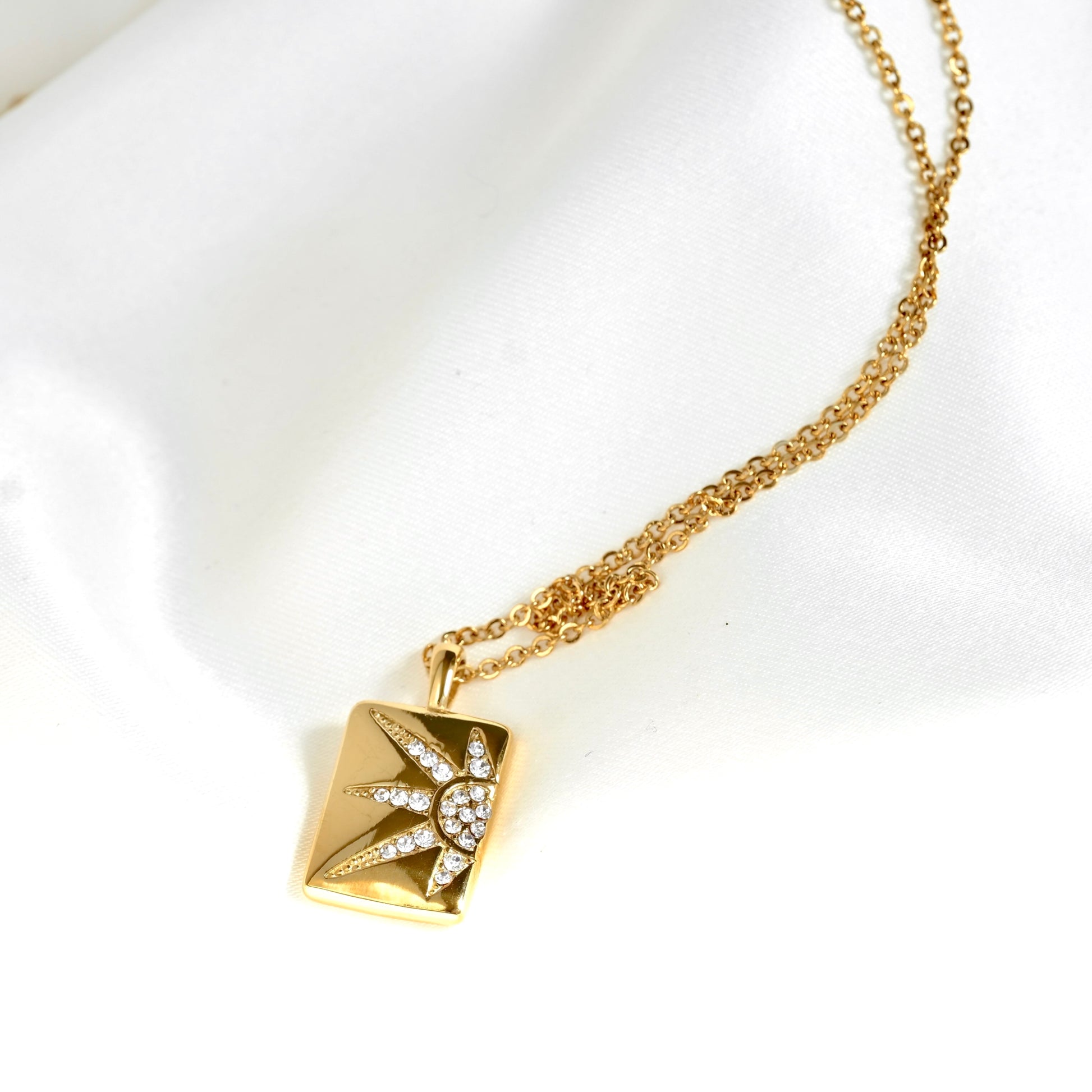 A gold sun pendant necklace featuring a square pendant with a sunburst design set with small clear stones, displayed on a white satin fabric background.