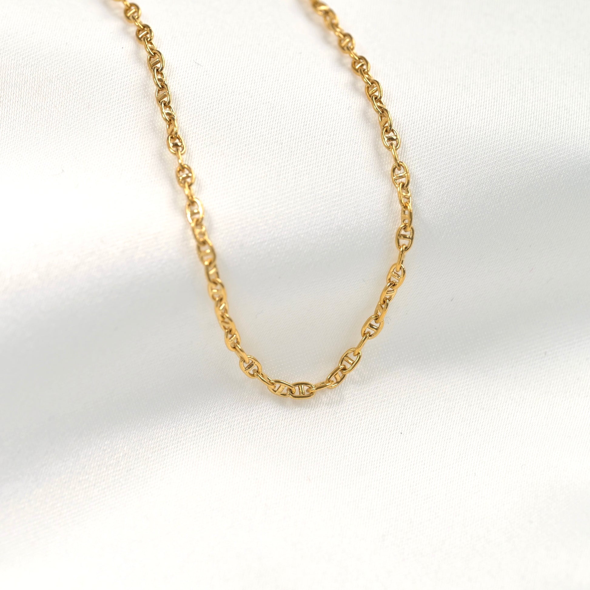 A delicate gold mariner necklace with an oval link design, displayed on a soft white fabric background.