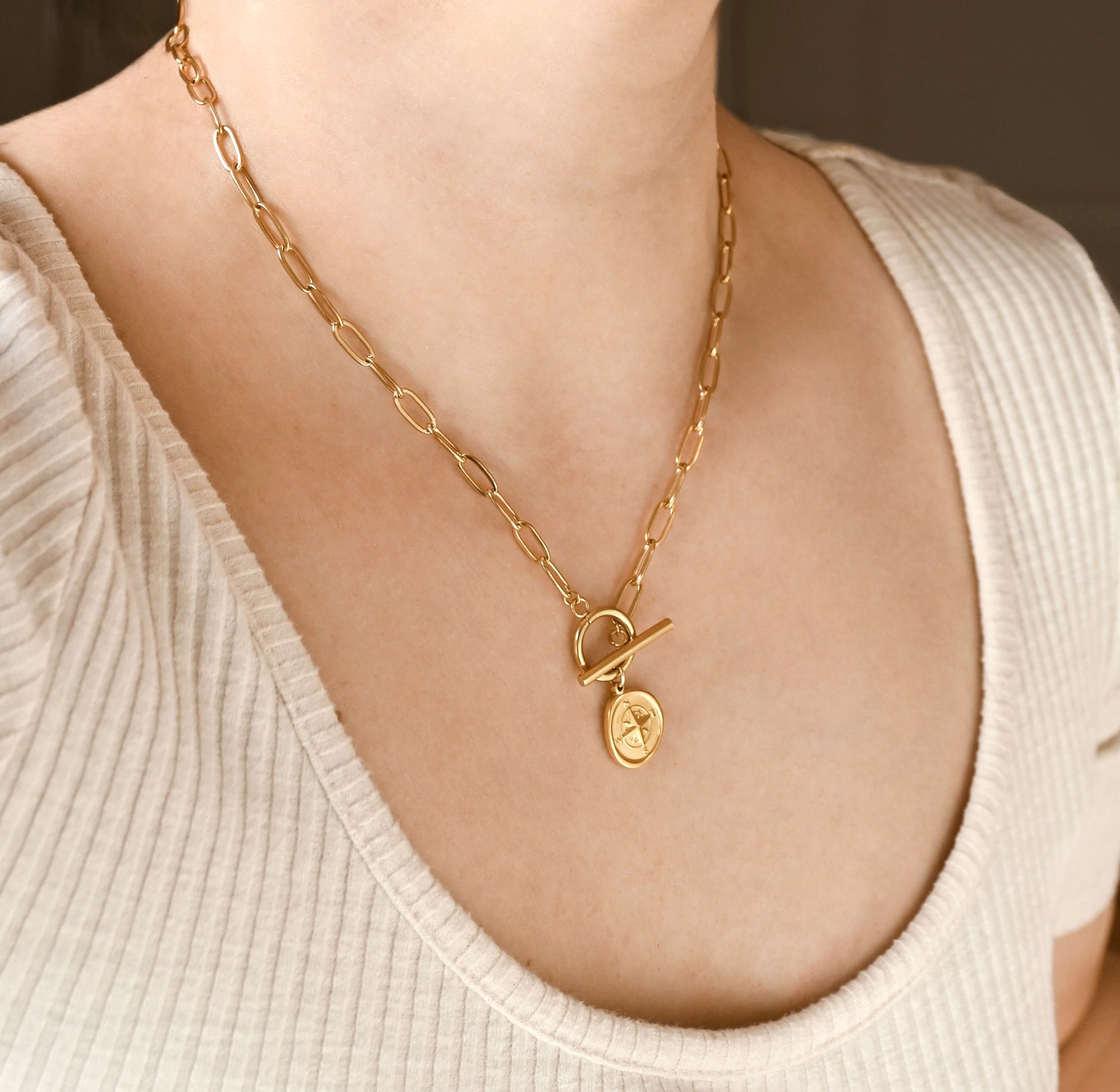 A close-up of a person wearing a gold chain necklace with a toggle clasp and a small oval pendant featuring a star design, styled with a cream ribbed top.