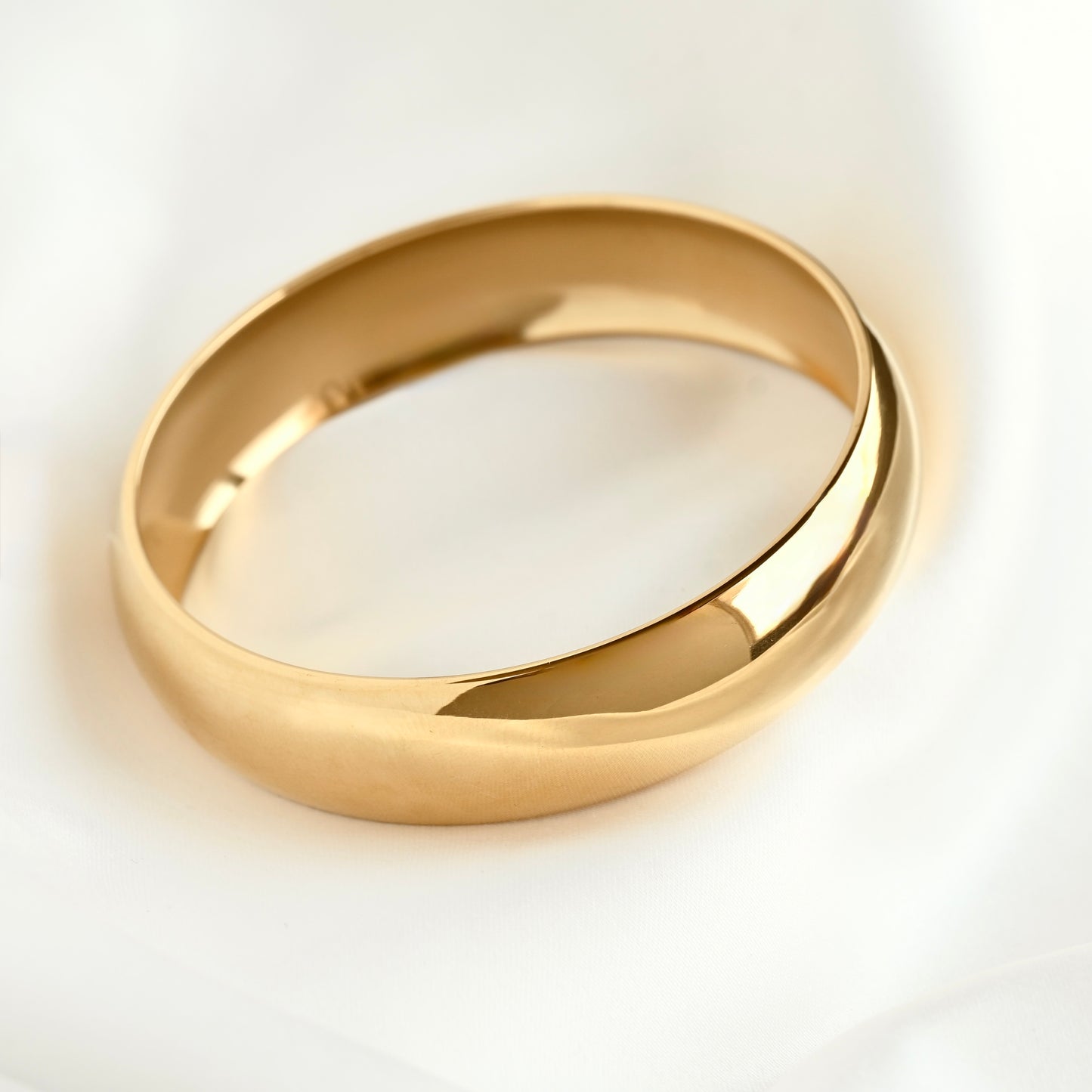 A polished thick gold bangle resting on a soft, white draped fabric background.