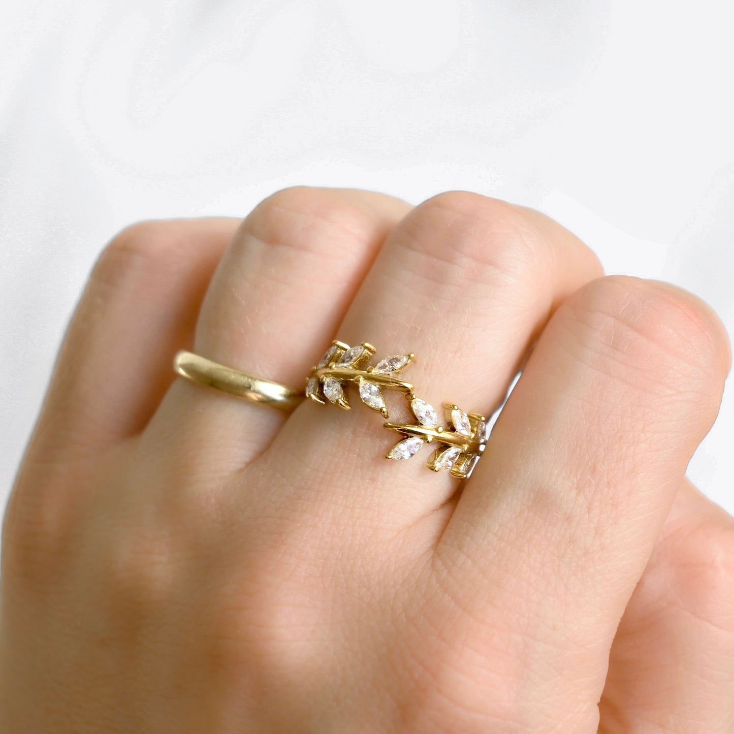 A close-up of a hand wearing a gold ring designed as a leafy branch with small clear stones, set against a soft white background.