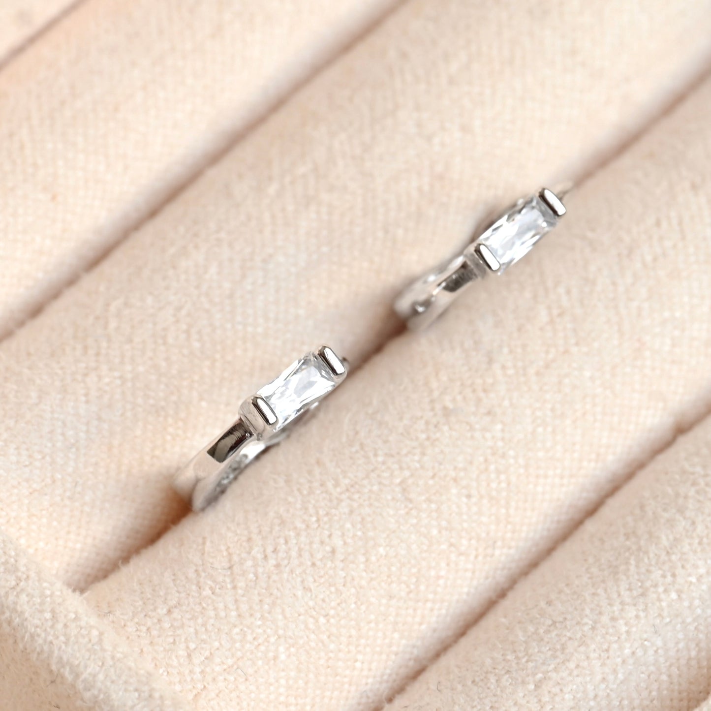 A pair of silver  huggie hoop earrings with rectangular clear gemstones, displayed on a beige textured ring stand.
