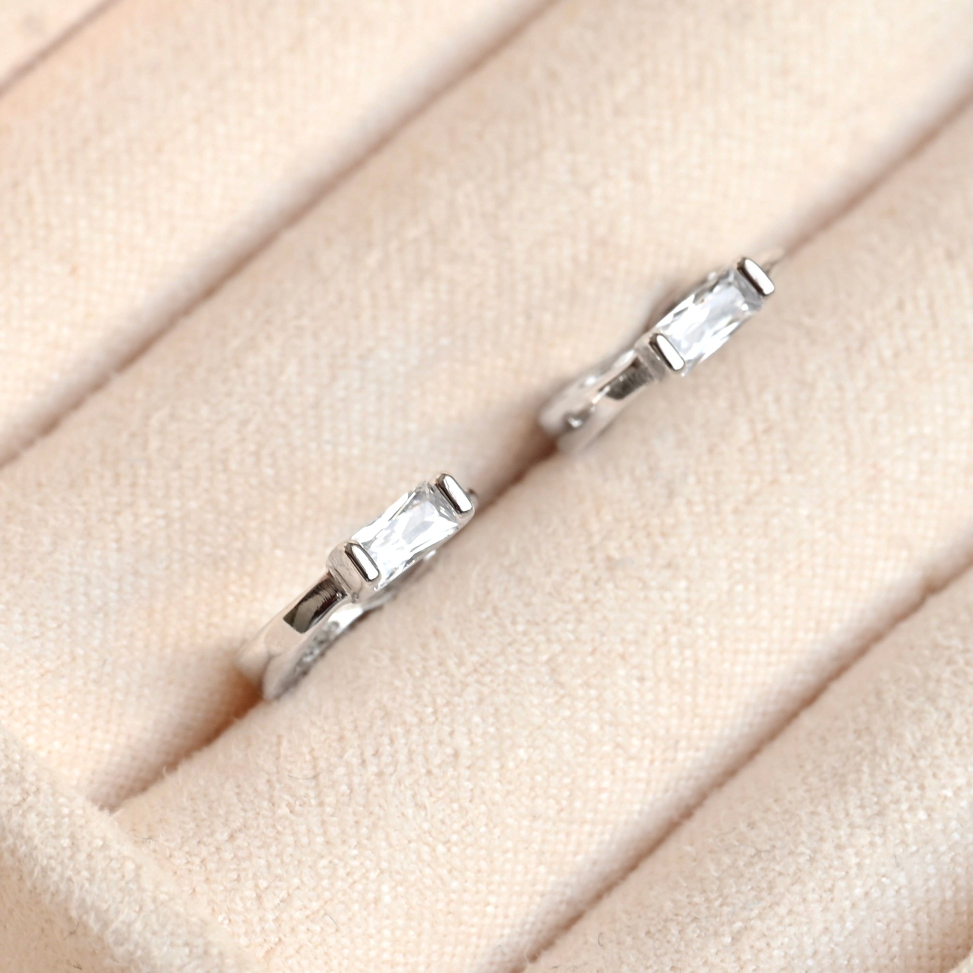 A pair of silver  huggie hoop earrings with rectangular clear gemstones, displayed on a beige textured ring stand.