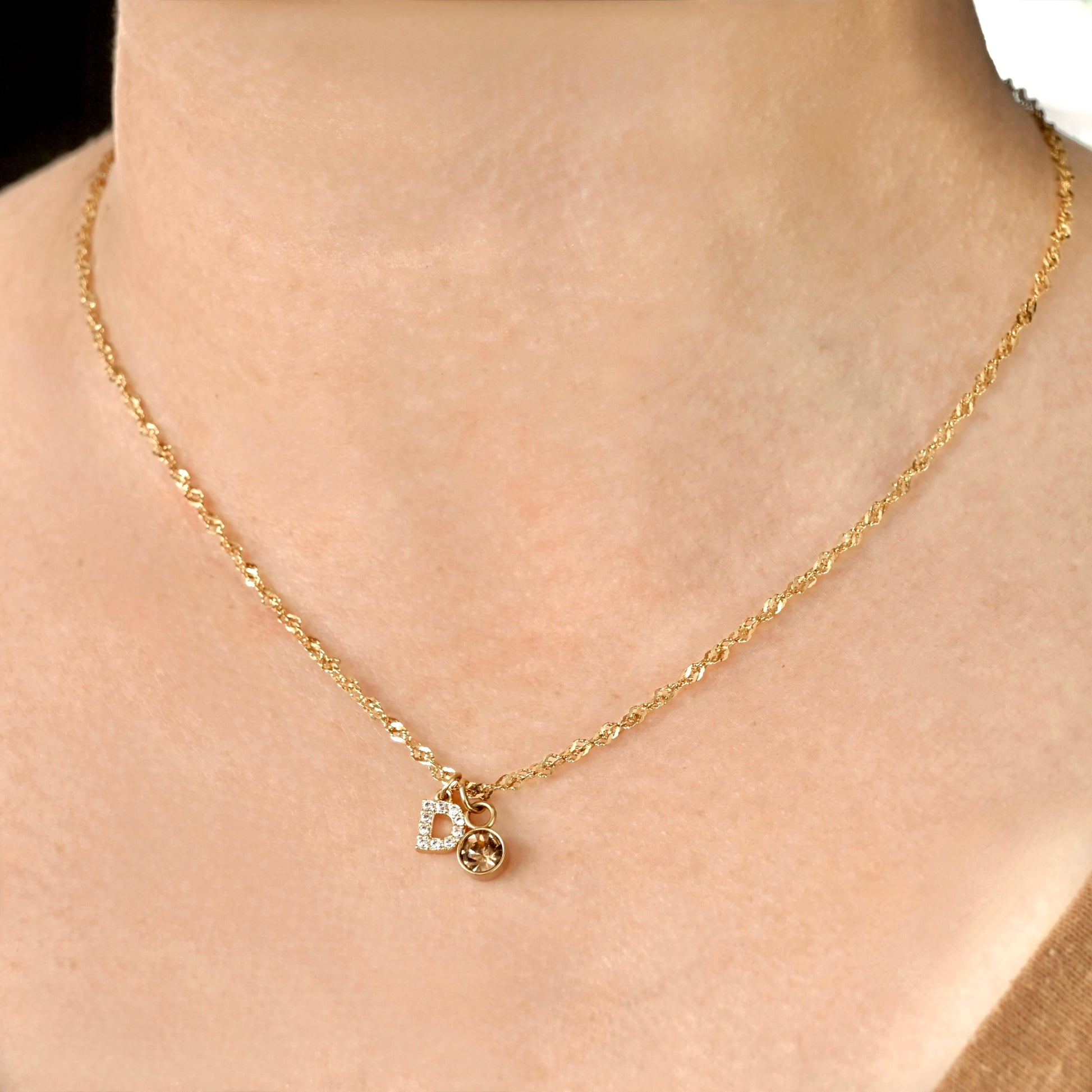 Gold twisted chain with a diamond 'D' pendant  and november birthstone on a person's neck against a neutral background