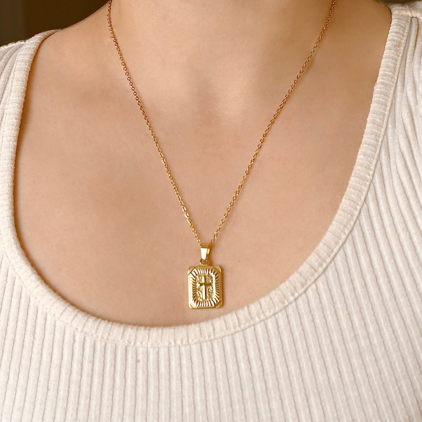Gold necklace with a cross tablet pendant with sunburst detail on a person wearing a beige top.