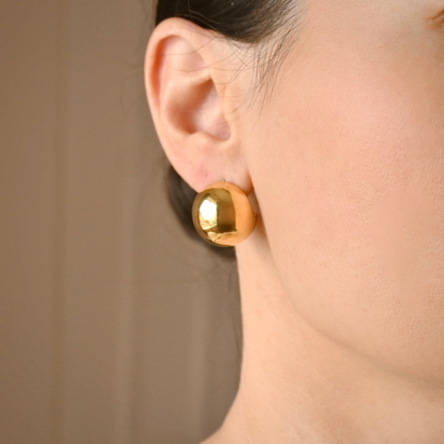 A close-up of a person's ear wearing a shiny gold spherical stud earring against a soft beige background.