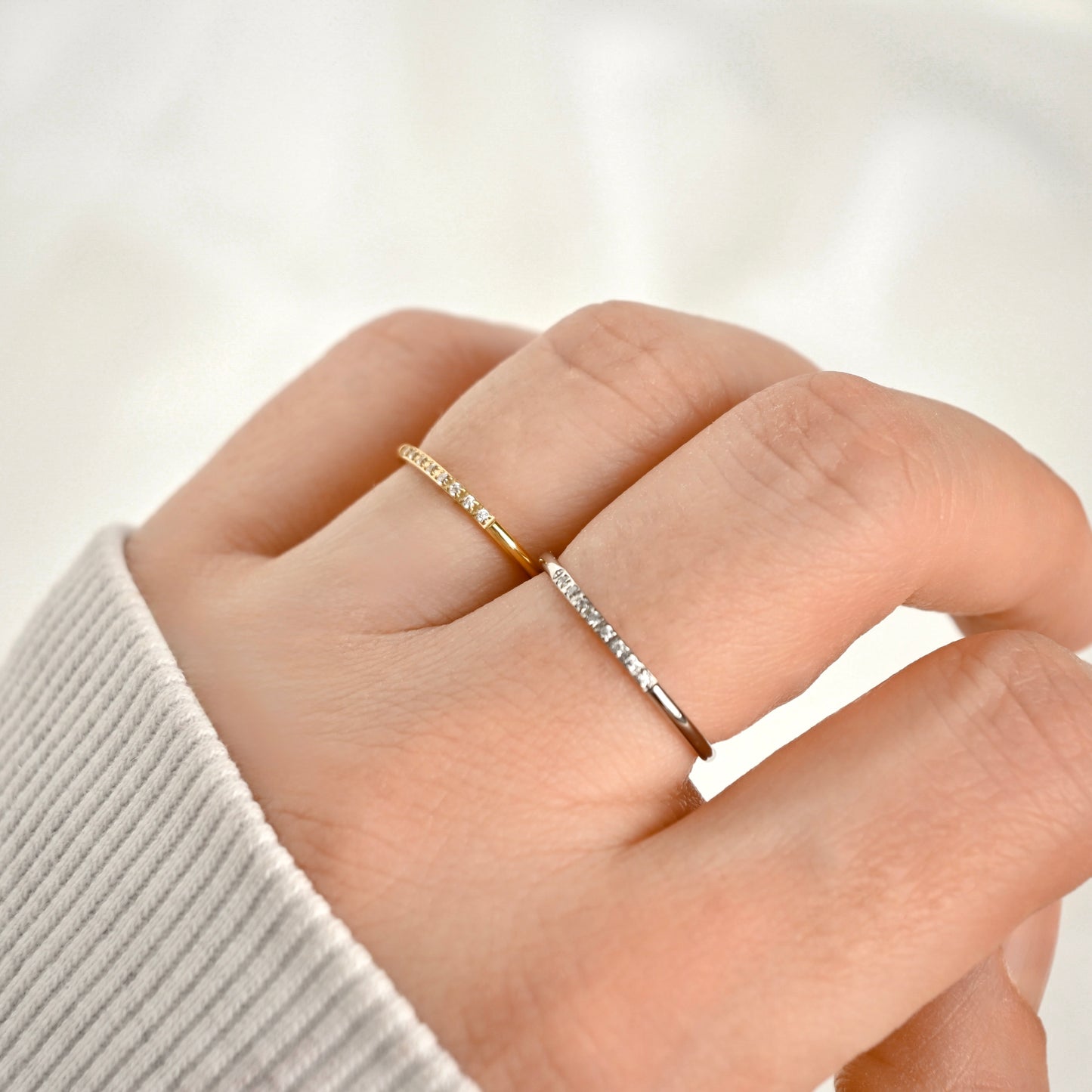 A close-up of a hand wearing two delicate rings: one gold and one silver band with tiny clear stones, against a soft white background.