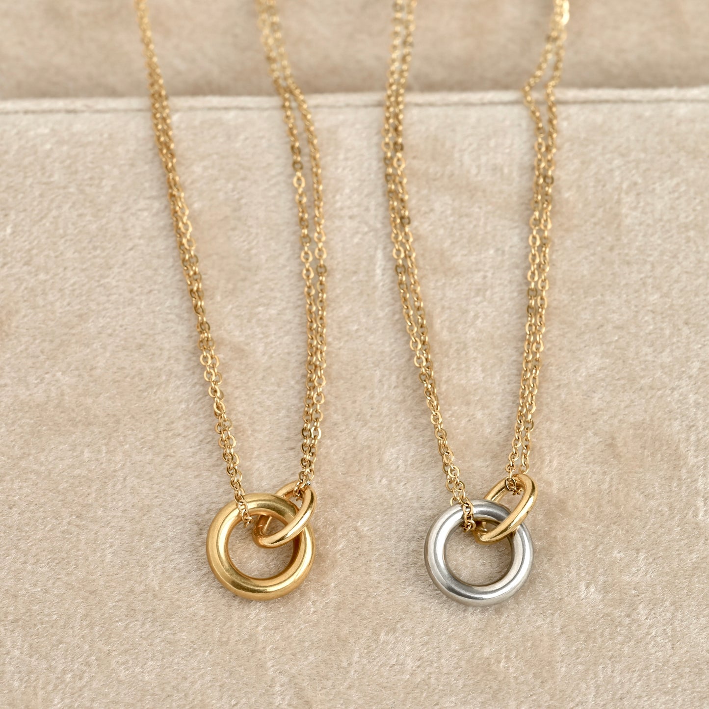 Two delicate chain necklaces with interlocking double-ring pendants displayed on a beige textured surface. One pendant is gold-toned and the other is silver-toned.