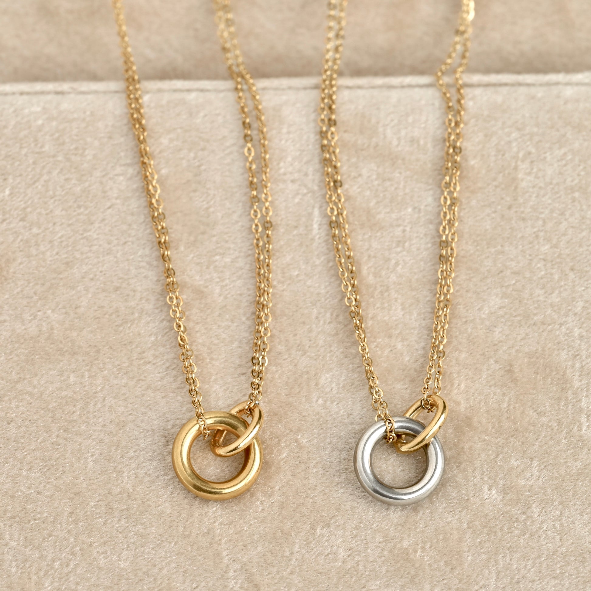 Two delicate chain necklaces with interlocking double-ring pendants displayed on a beige textured surface. One pendant is gold-toned and the other is silver-toned.