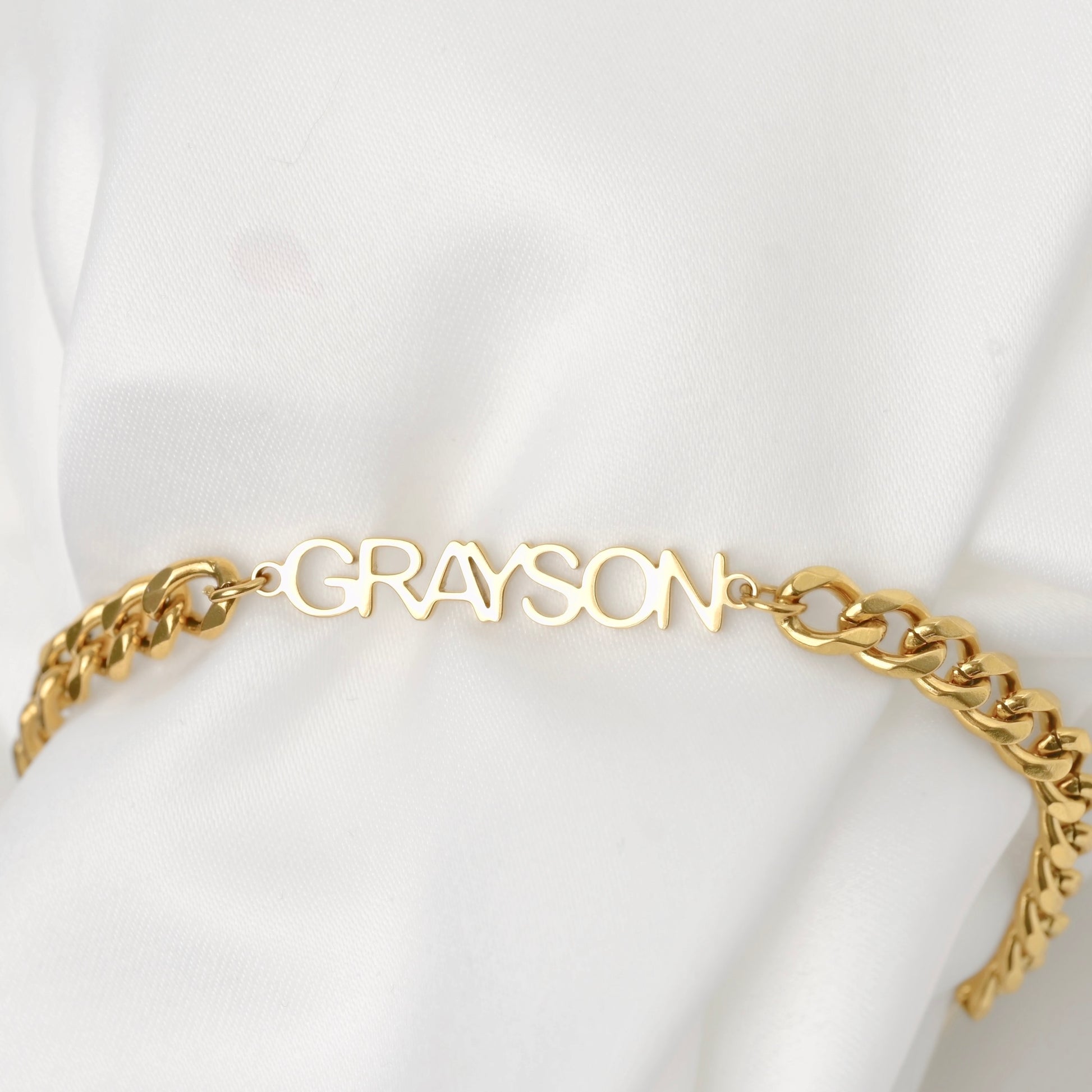 A thick gold 5mm cuban chain bracelet with a custom nameplate spelling 'GRAYSON', displayed on a soft white fabric background.