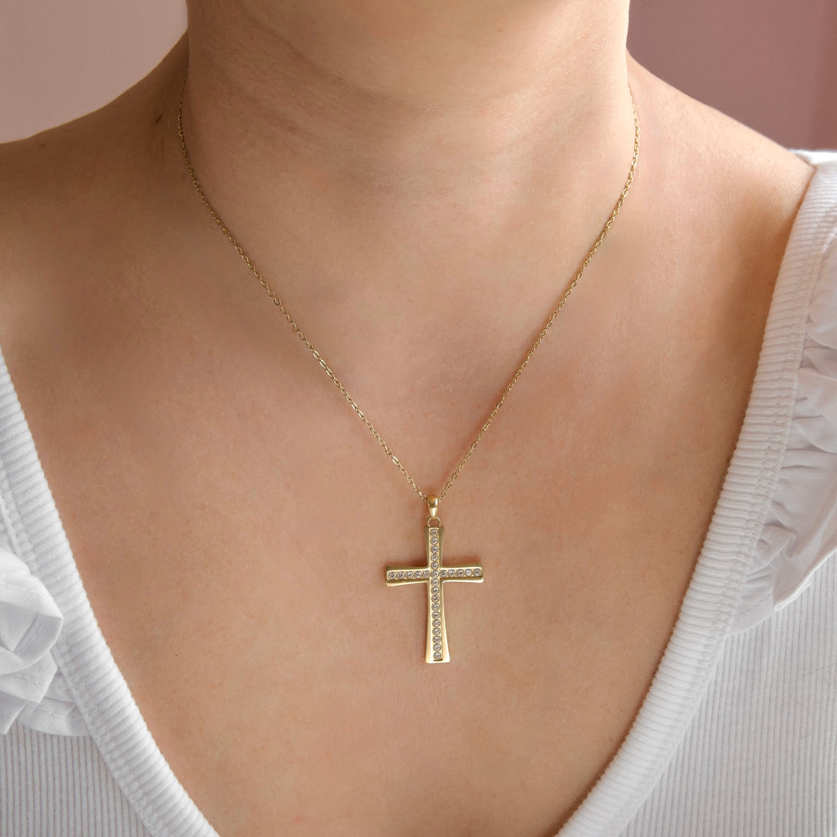 A gold cross pendant necklace with small clear stones, worn on a model's neck over a white top.