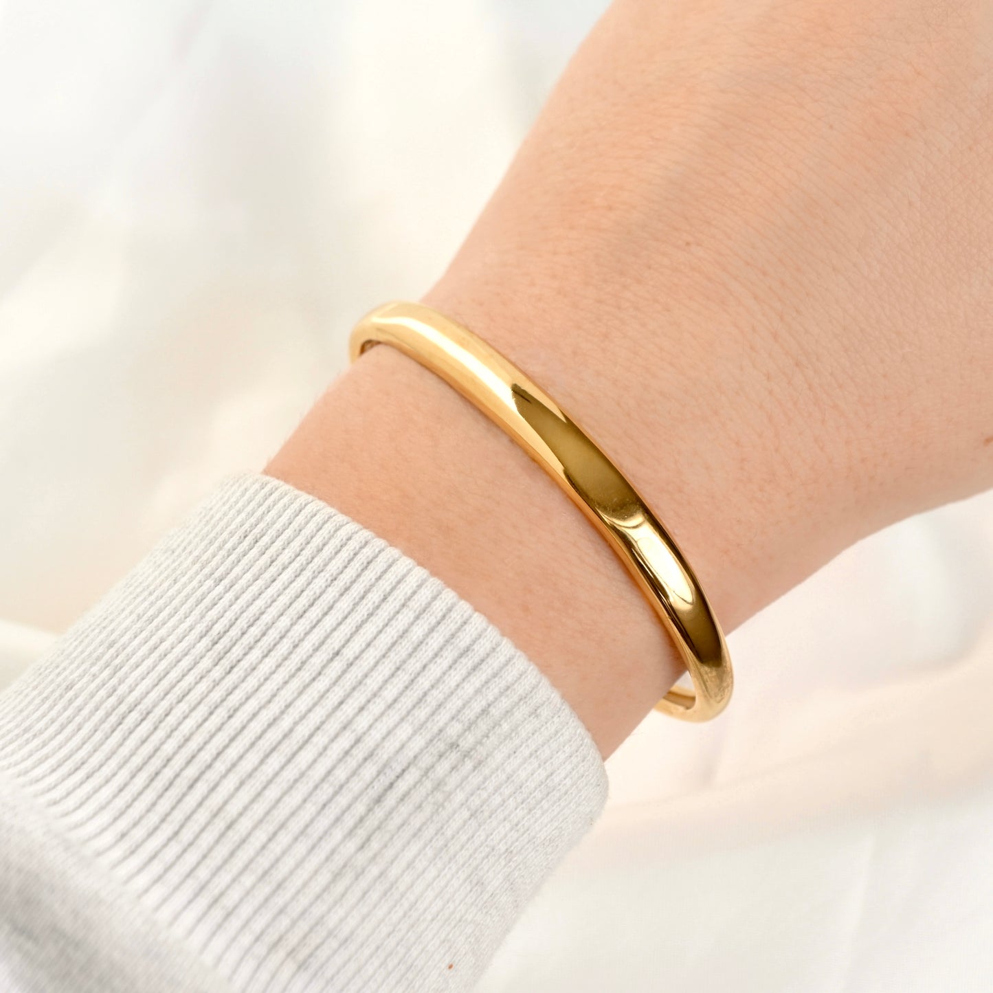 A polished gold cuff bracelet worn on a person's wrist, shown against a soft white background with a light gray ribbed sleeve visible.
