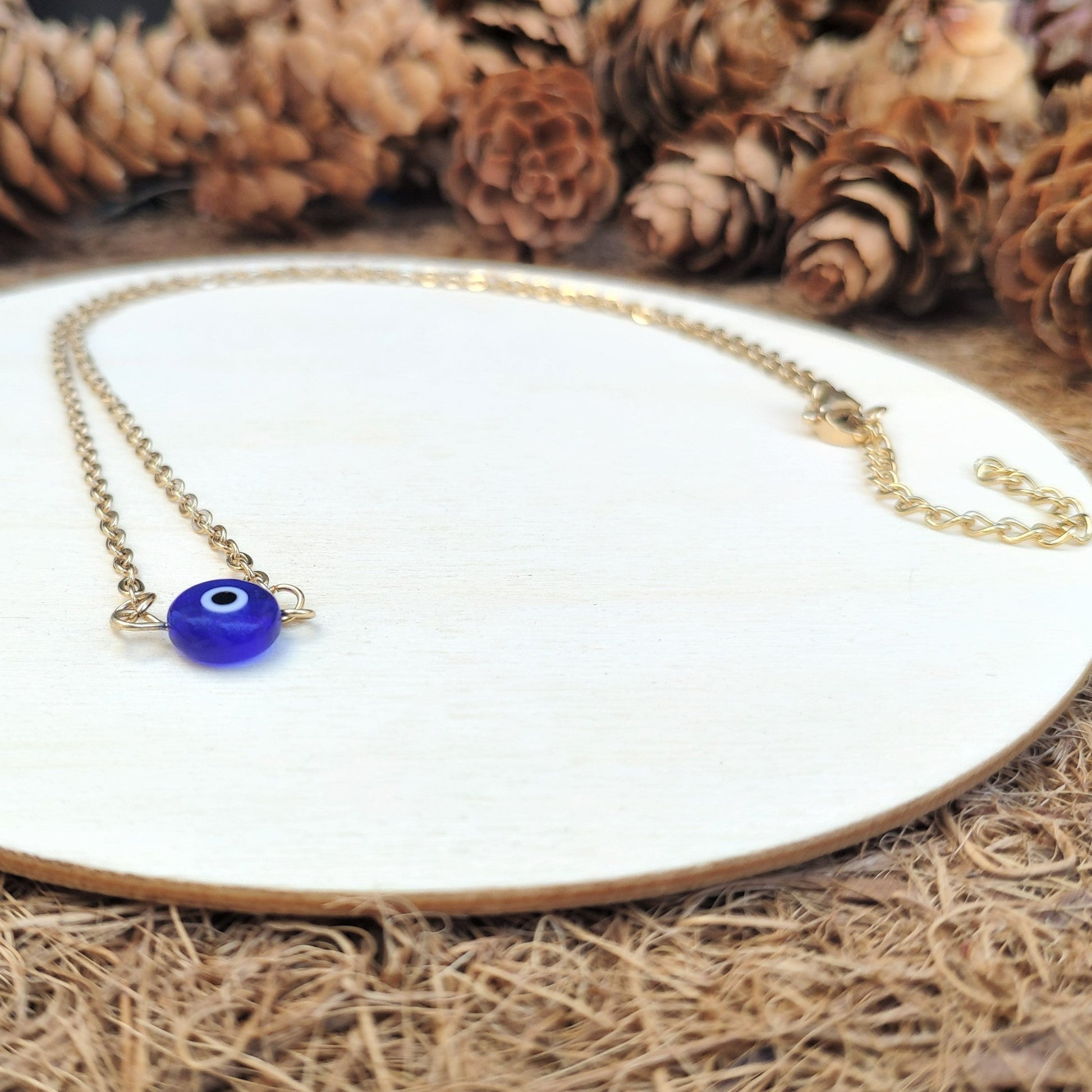 Necklace with a blue evil eye pendant on a white plate surrounded by pine cones.