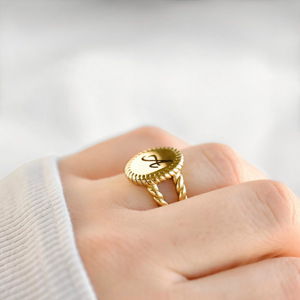 A gold ring with a twisted rope band and a round top engraved with a black monogram, worn on a person's finger.