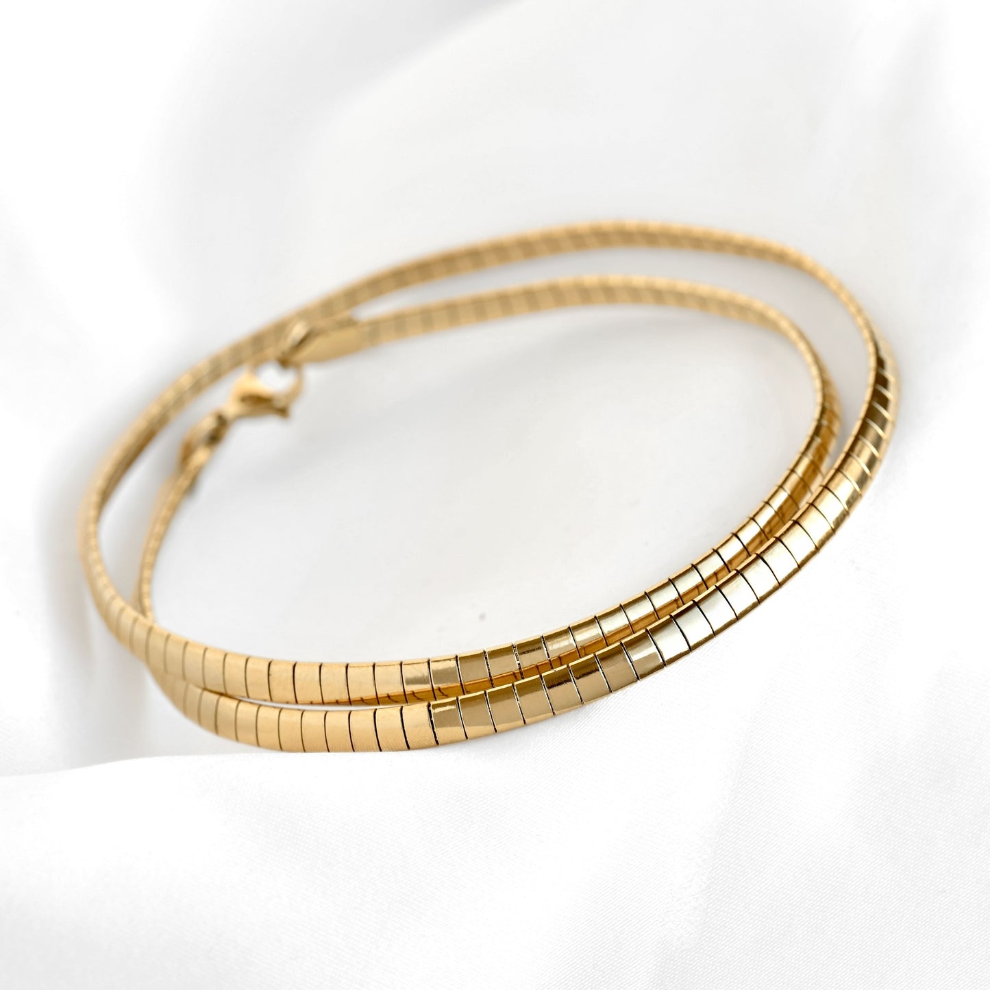 A gold-toned  omega chain necklace with a lobster clasp, resting on a white fabric background.
