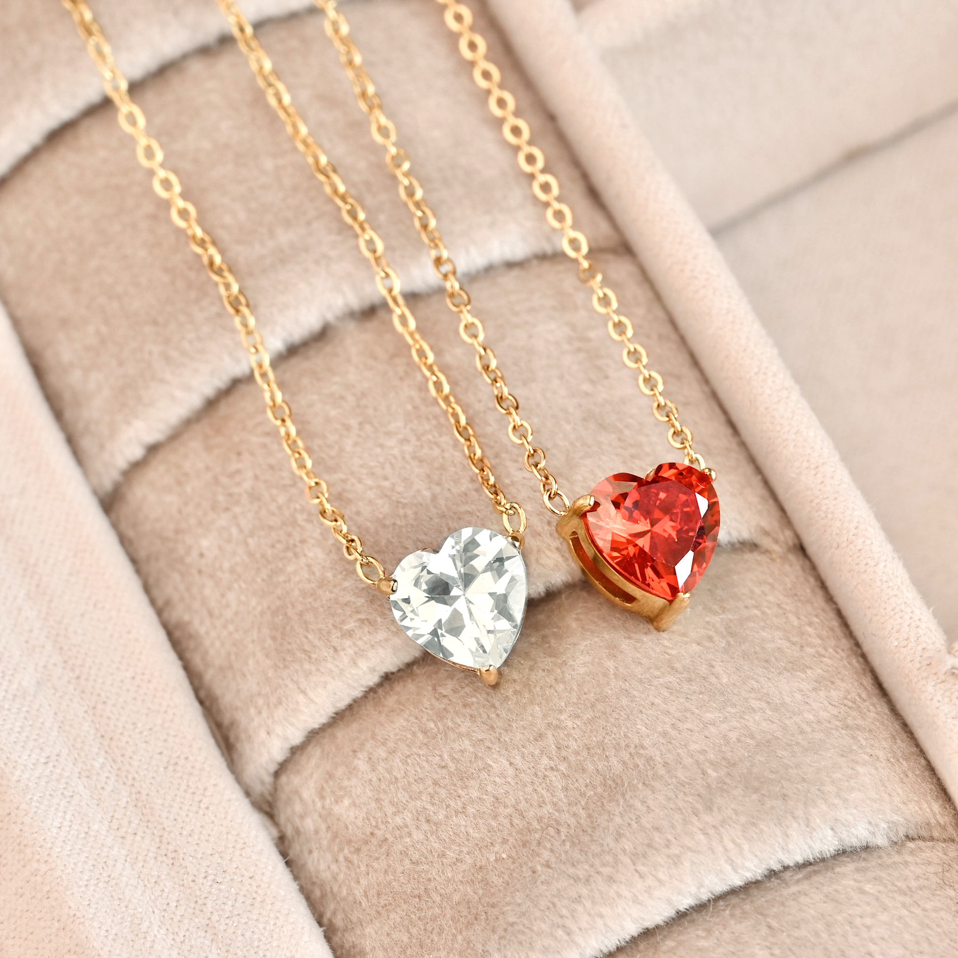Two gold necklaces with heart-shaped pendants, one clear and one red, on a soft fabric background.