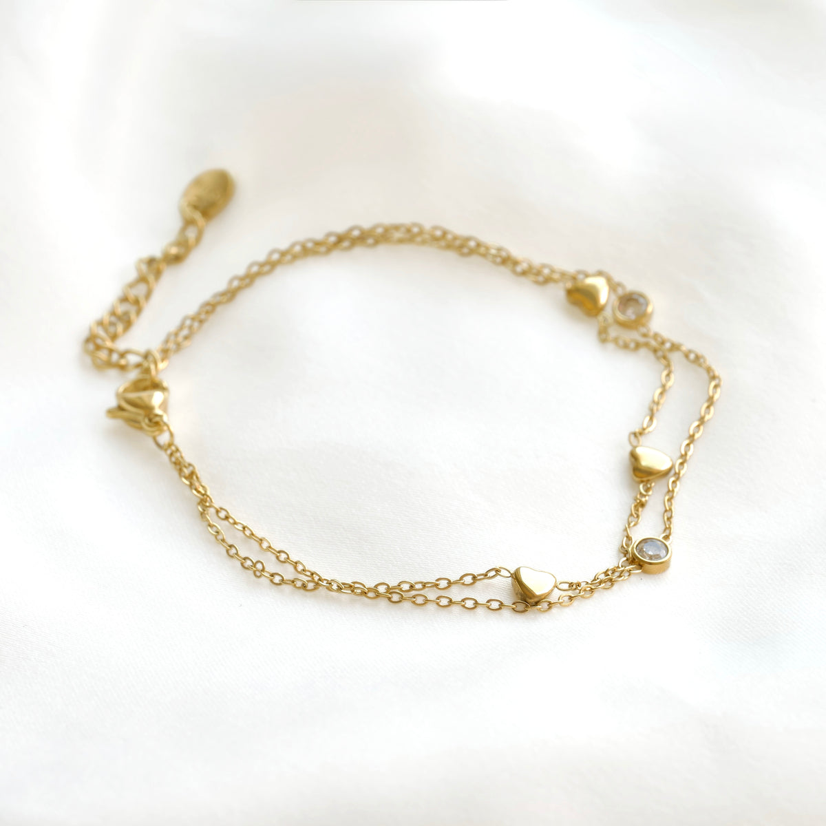 A delicate gold chain bracelet featuring small heart charms and two clear round bezel-set stones , displayed on a soft white fabric background.