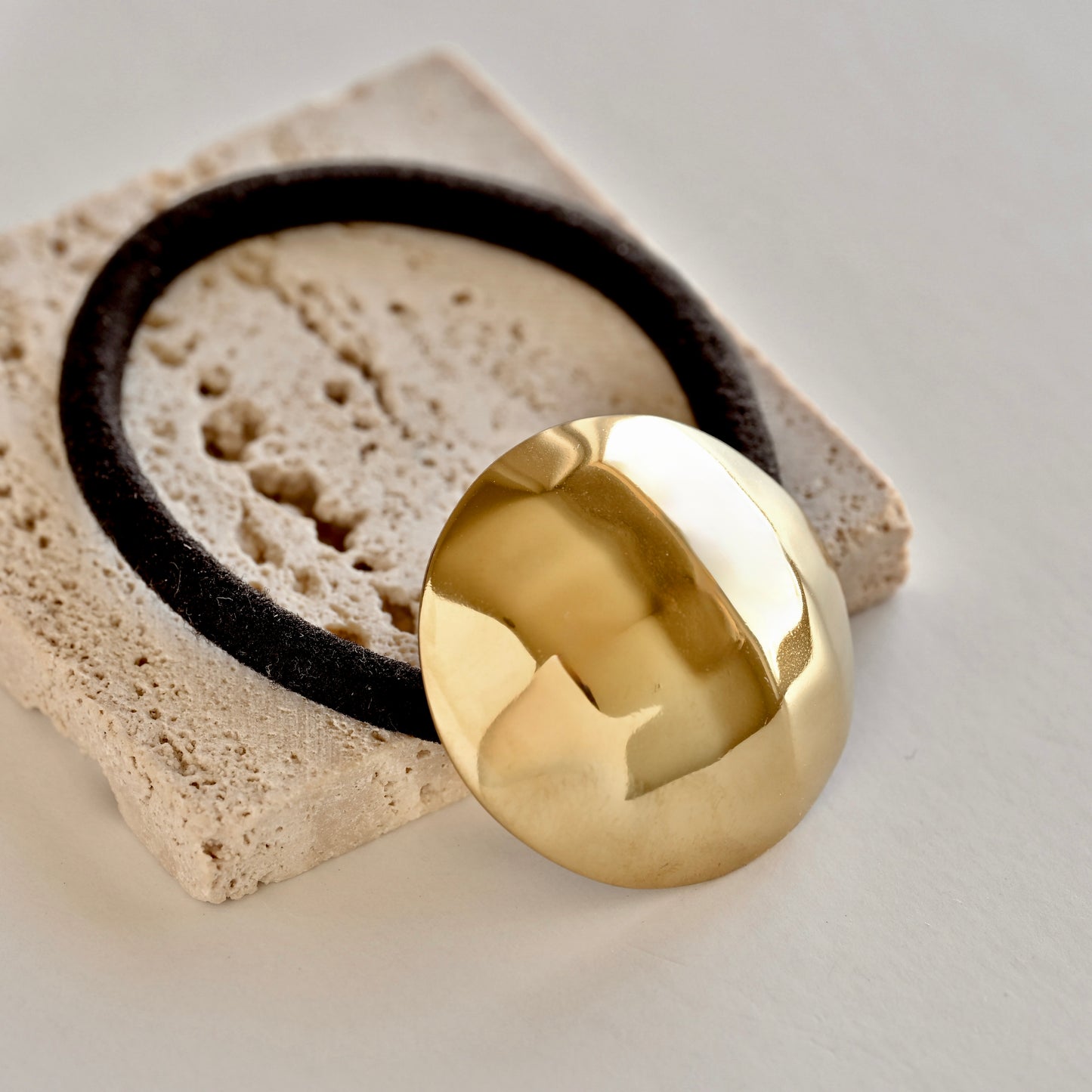 A polished, highly reflective gold circular disc or medallion resting on a textured beige stone tile, with a black circular ring partially visible behind it.
