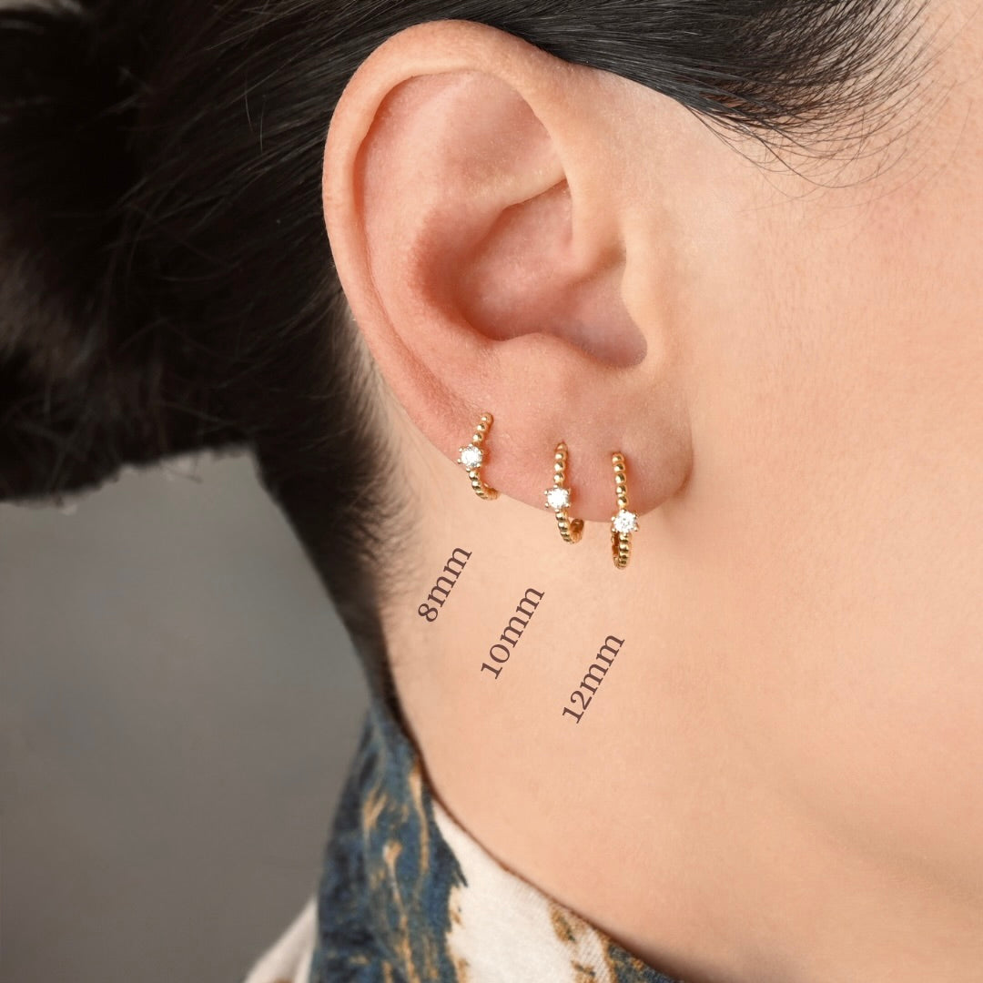 Close-up of a person's ear wearing three small gold hoop earrings with tiny clear stones, with size labels 8mm, 10mm, and 12mm overlaid near the earrings.
