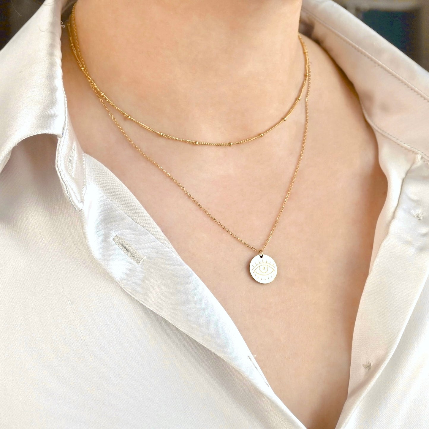 Angled front view of person wearing a layered gold evil eye necklace with a round eye engraved pendant  reflecting light 