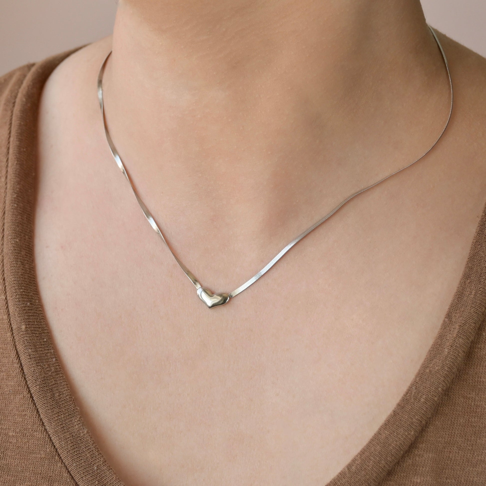 Silver snake chain necklace with a heart pendant on a person wearing a brown top.