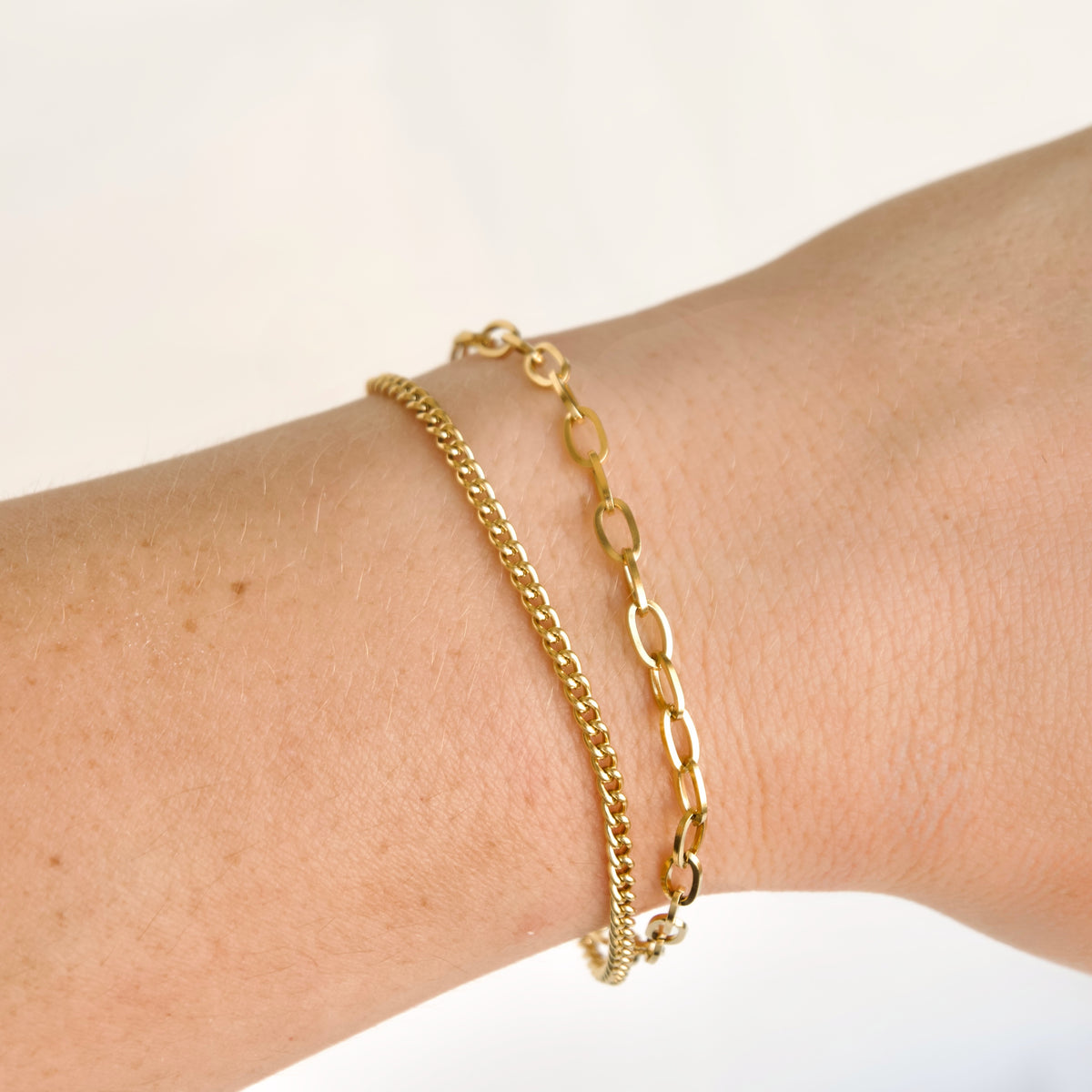 Double strand gold chain bracelet with a fine curb chain and a shiny oval link chain worn on a wrist.