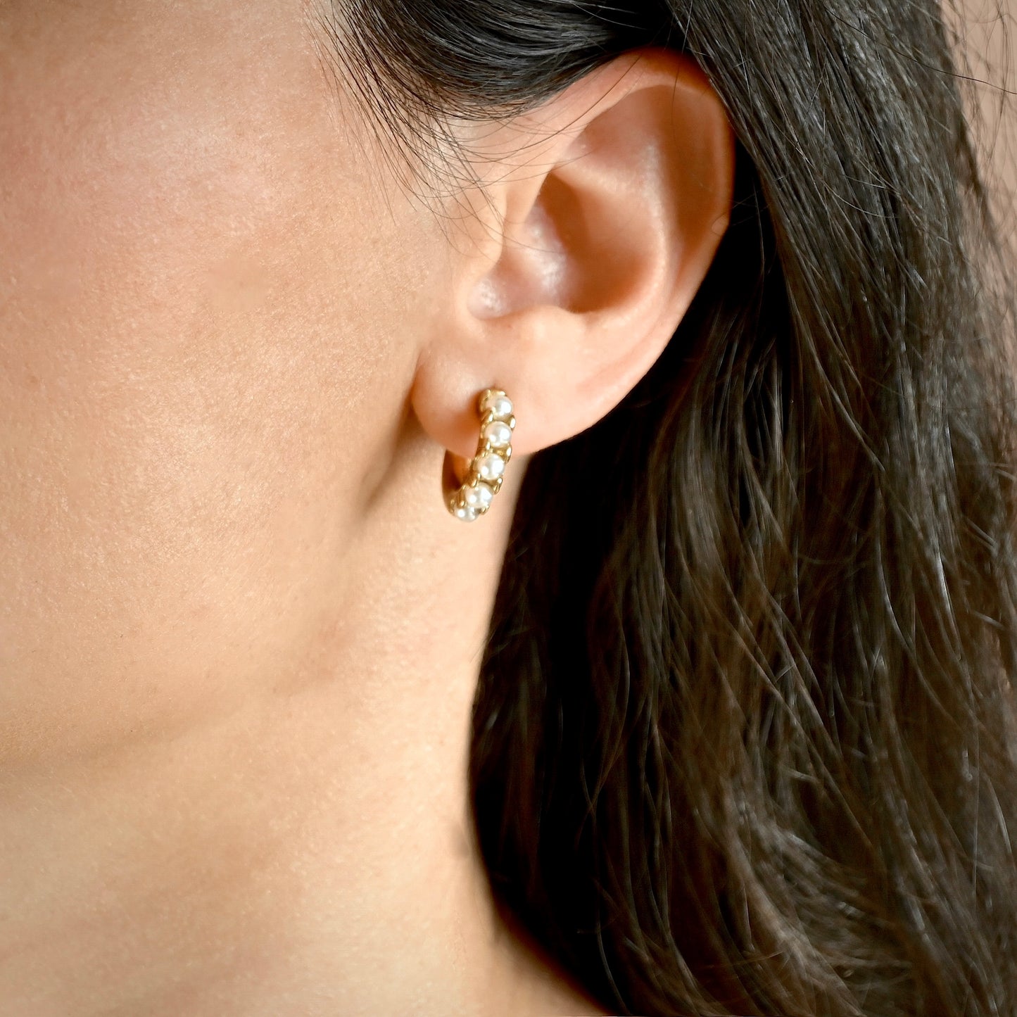 A close-up of a person's ear wearing a small gold huggie hoop earring adorned with cwhite pearls.
