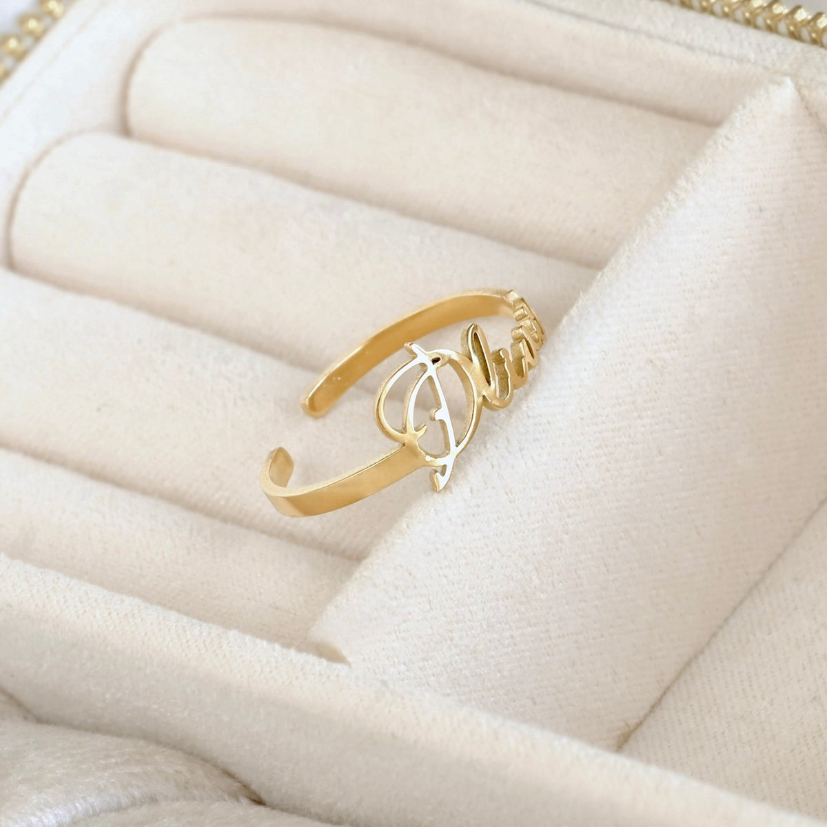 A gold-plated open cuff ring featuring a central script monogram design, displayed on a cream-colored velvet jewelry box.