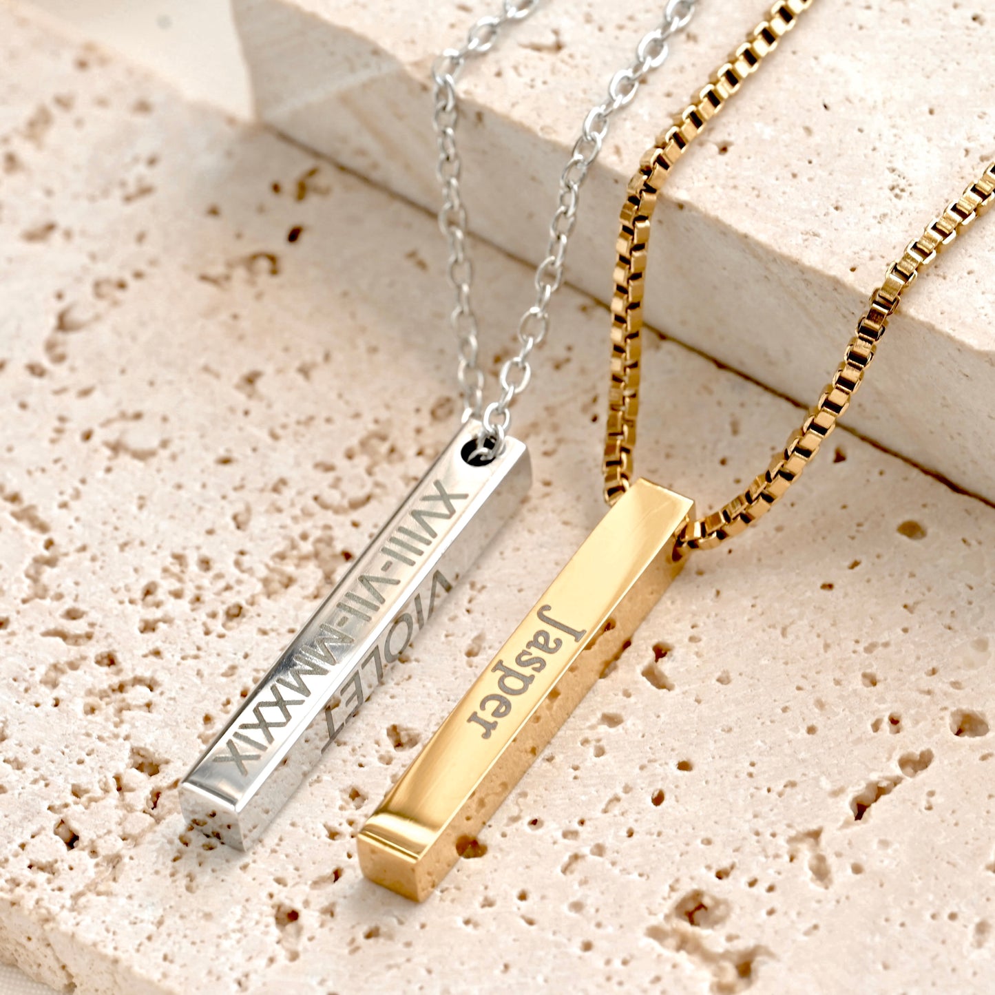Gold and silver bar necklaces , one with cable chain and other box chain, on a textured beige stone