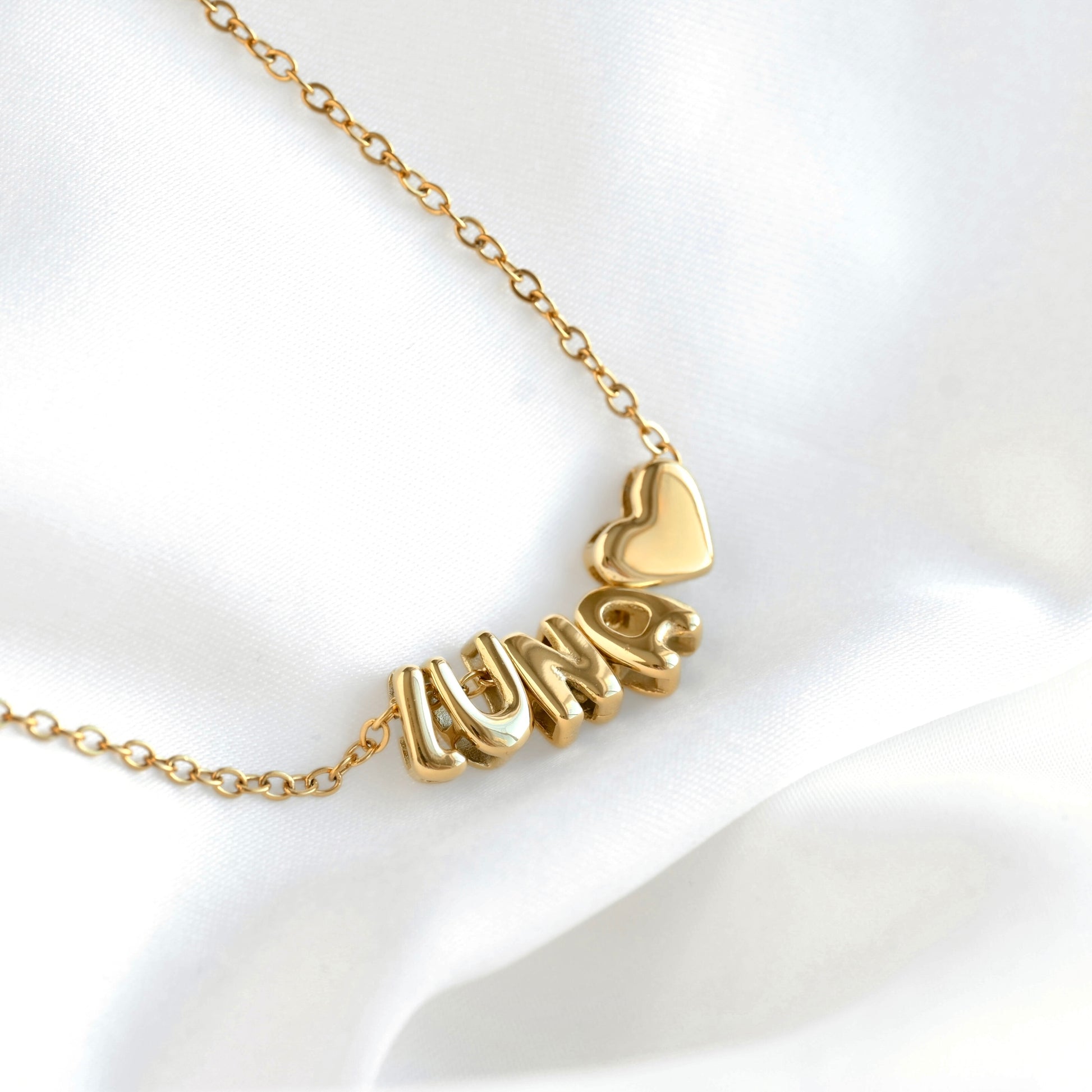 A gold chain necklace featuring a personalized nameplate pendant spelling 'LUNA♡' in bubble like small letter charms, with a small heart charm, displayed on a soft white satin fabric background.