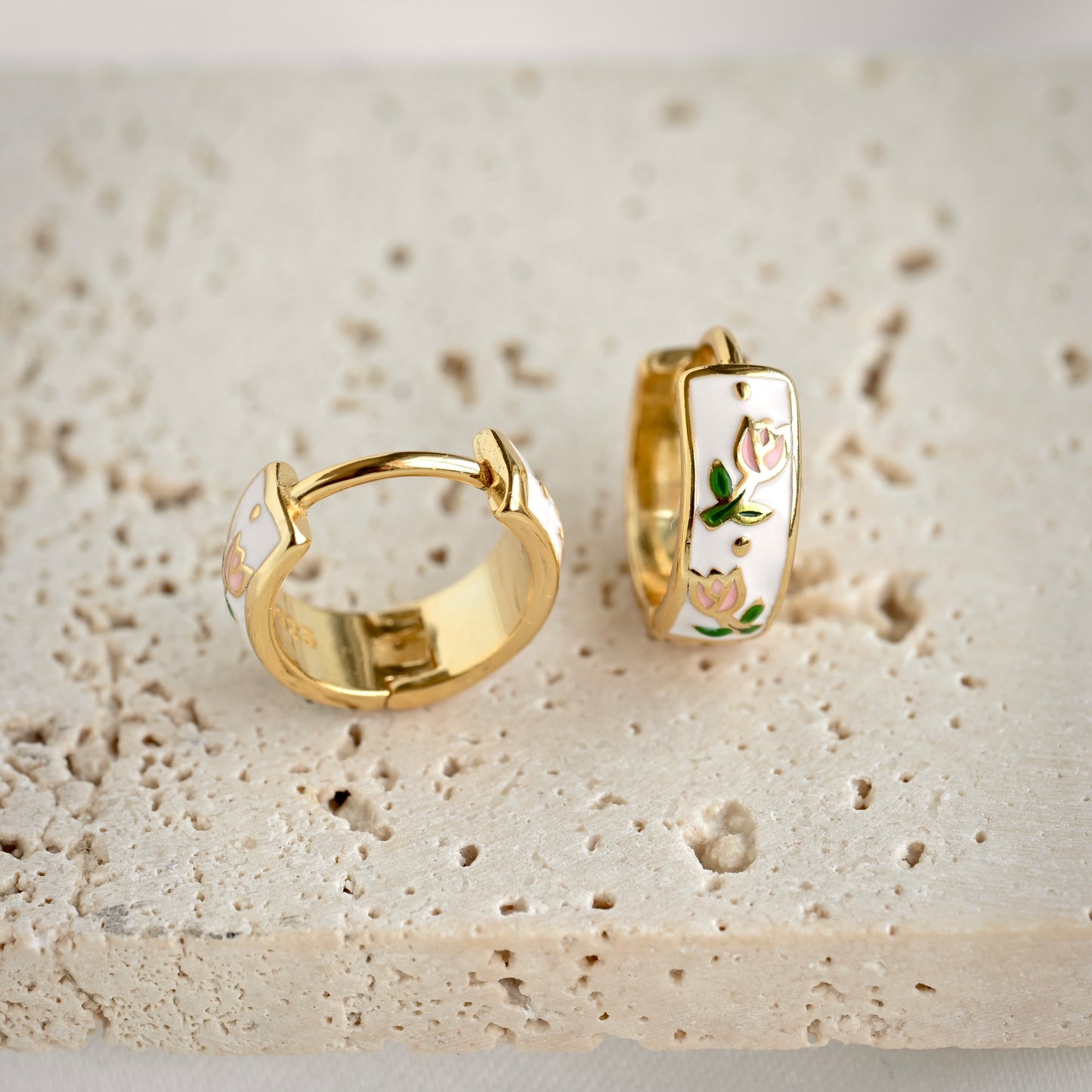 A pair of gold huggie hoop earrings with white enamel inlay and delicate pink floral designs, displayed on a textured beige stone surface.