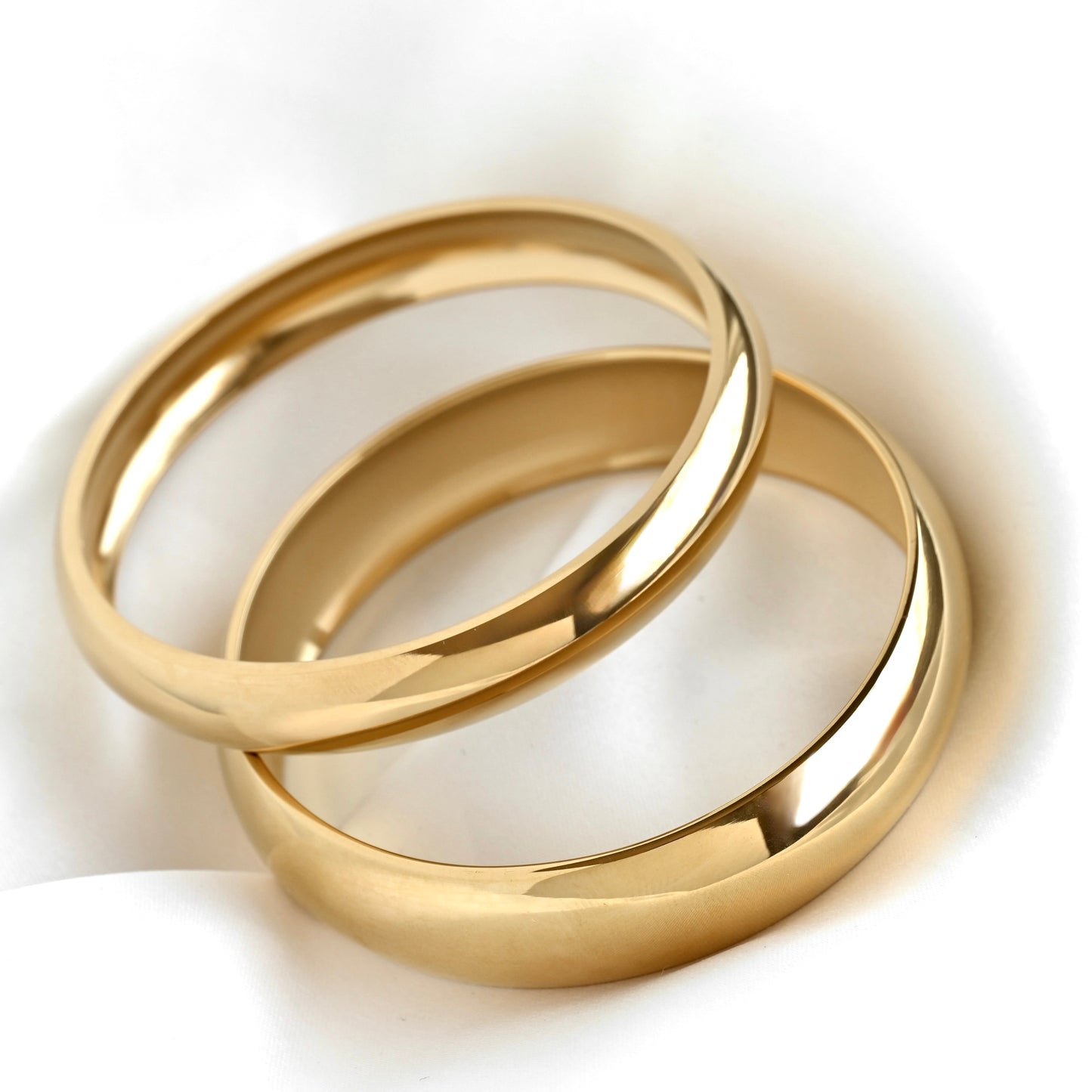 Two polished gold bangles, one thin and one wide, stacked together on a soft white fabric background.