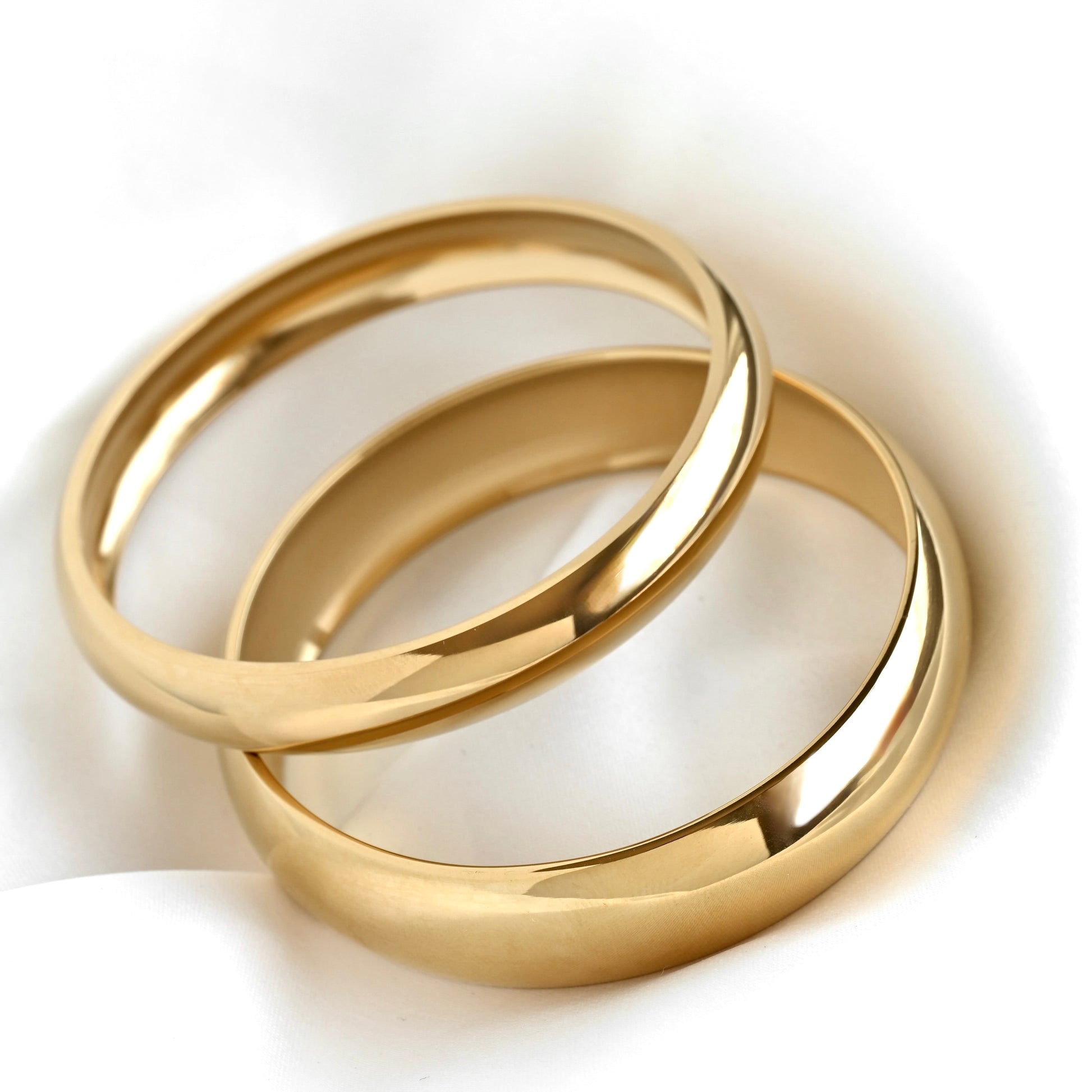 Two polished gold bangles, one thin and one wide, stacked together on a soft white fabric background.