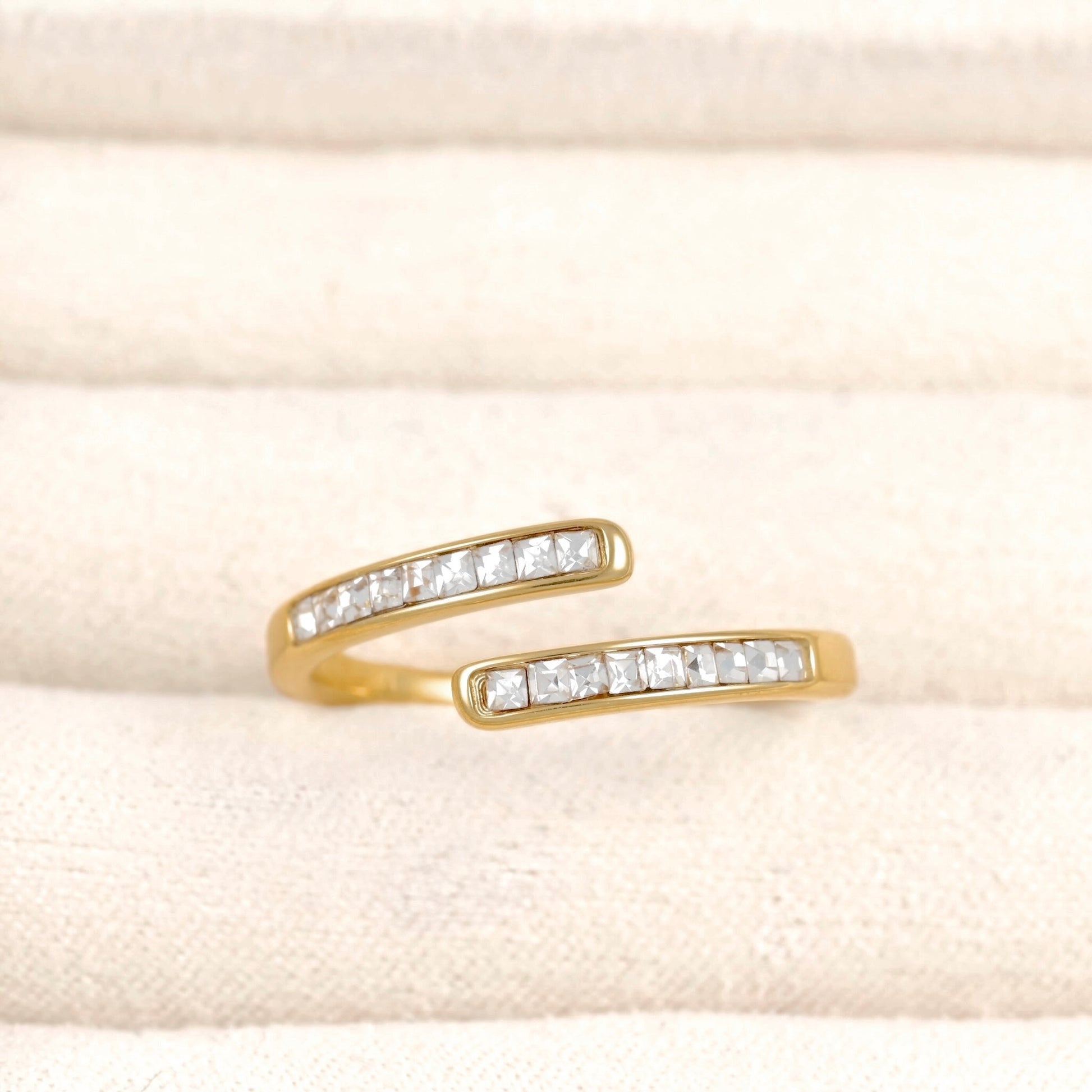 A gold open-band ring featuring a channel-set row of clear rectangular gemstones on each band, displayed on a textured cream fabric background.
