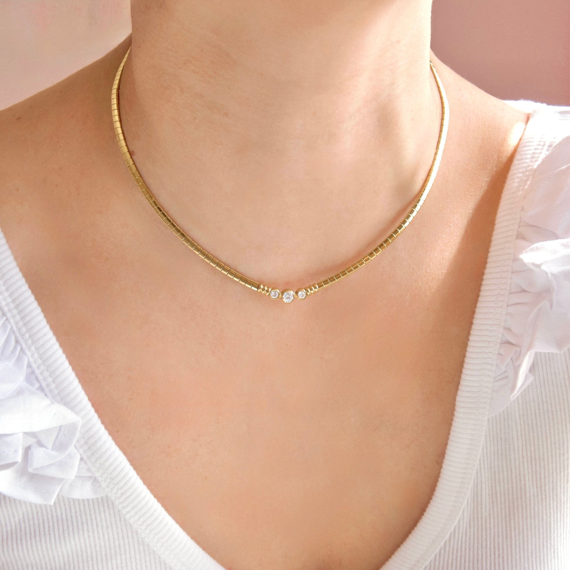 A close-up of a model wearing a delicate gold chain necklace with three small clear rhinestones at the center, styled with a white ruffled top.