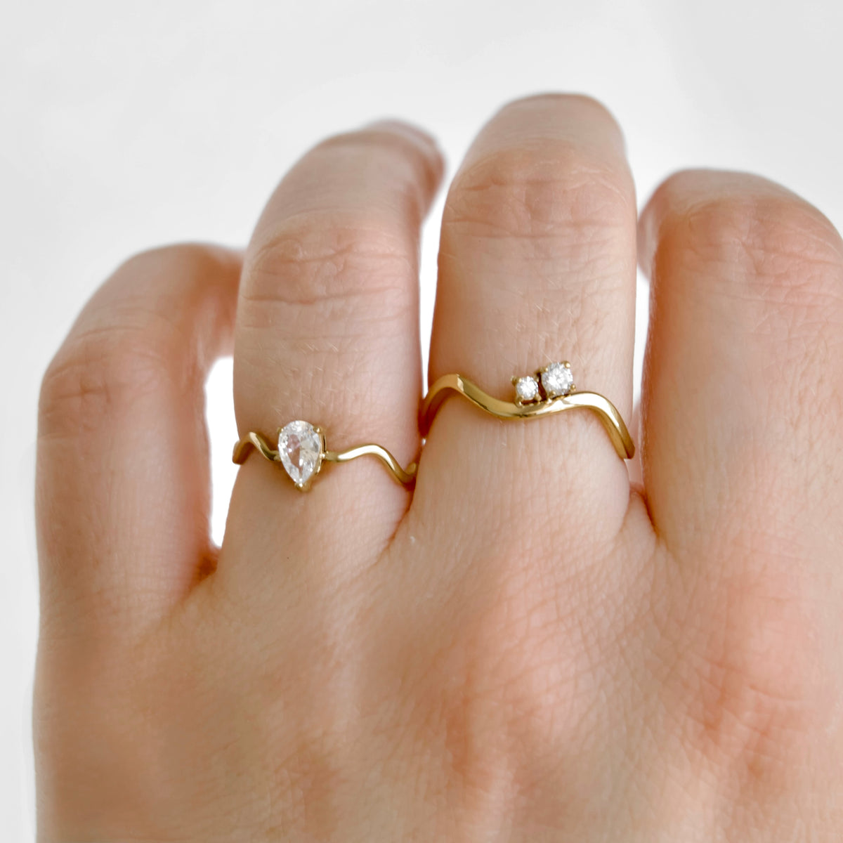 A close-up of a hand wearing two gold rings. One ring features a pear-cut clear gemstone on a wavy band, and the other features two small round clear gemstones on a wavy band.