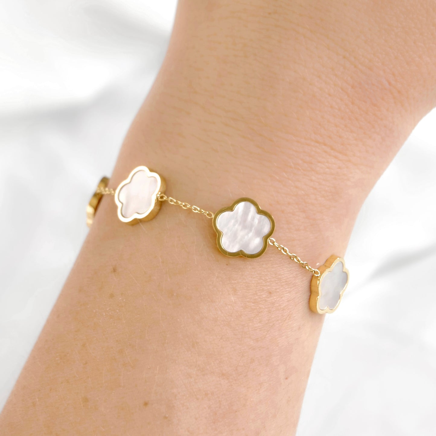 A delicate gold chain bracelet with three mother-of-pearl clover-shaped charms, shown worn on a person's wrist against a soft white background.