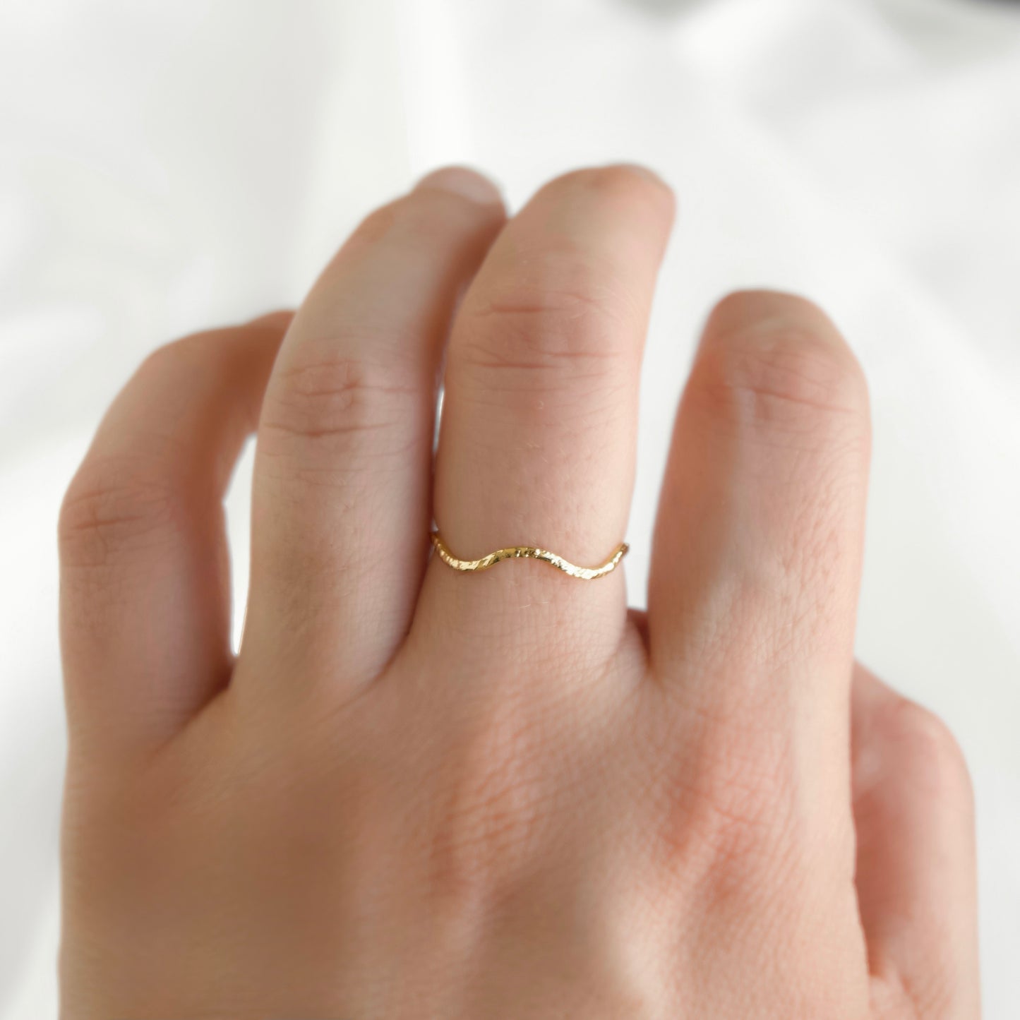 A delicate, thin gold ring with a wavy, undulating design worn on a person's ring finger, shown against a soft, light-colored fabric background.