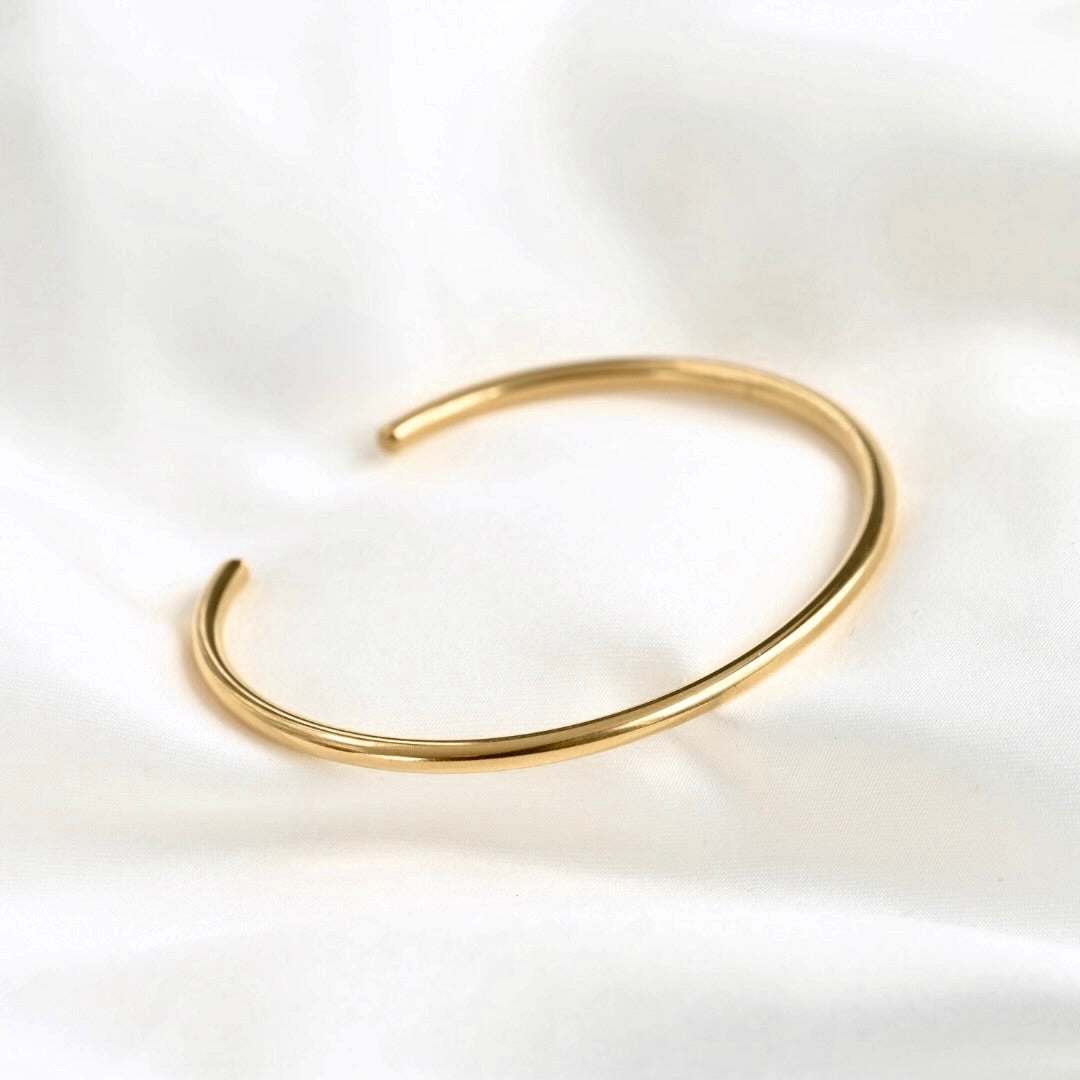 A minimalist gold open claw cuff bracelet resting on a soft, white draped fabric background.