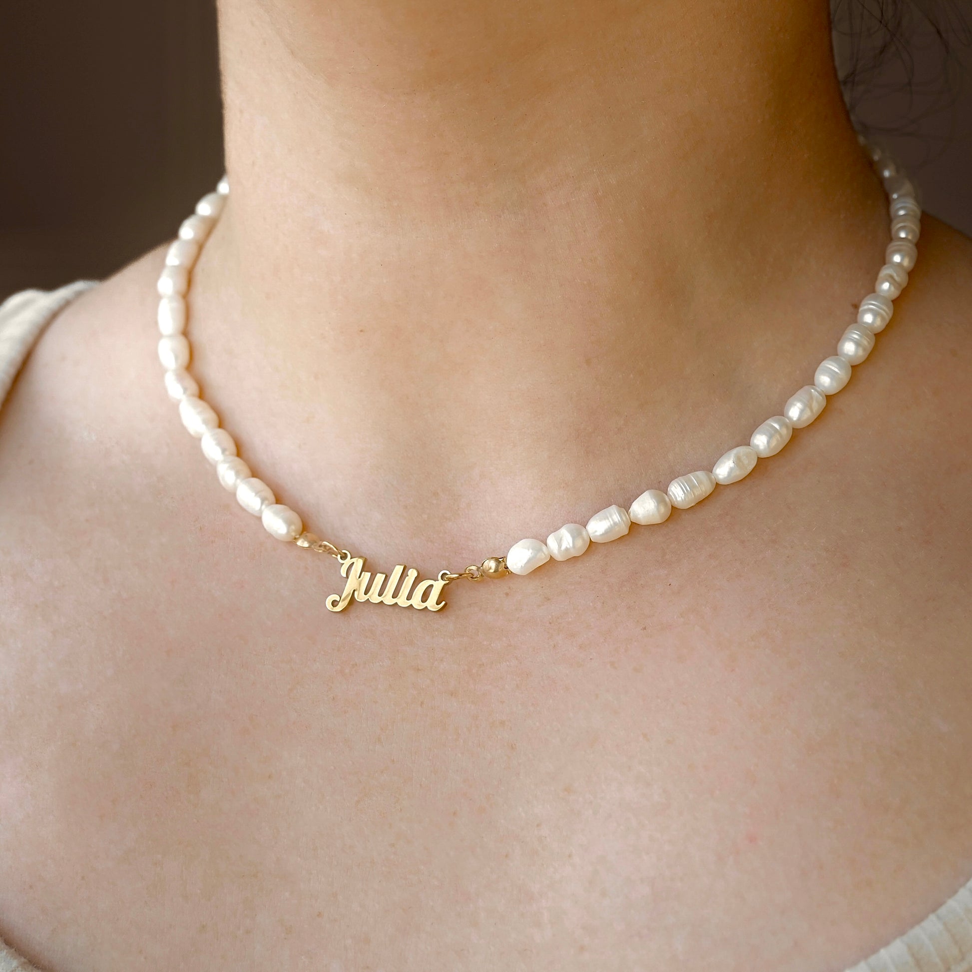 A close-up of a person's neck and collarbone wearing a personalized baroque pearl necklace with a gold script nameplate spelling 'Julia'.