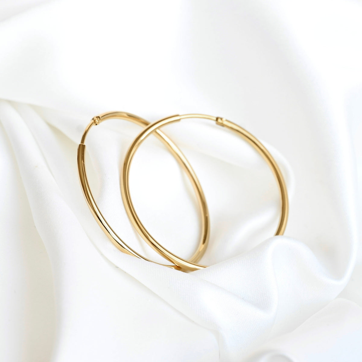 Two thin gold endless hoop earrings resting on draped white satin fabric.